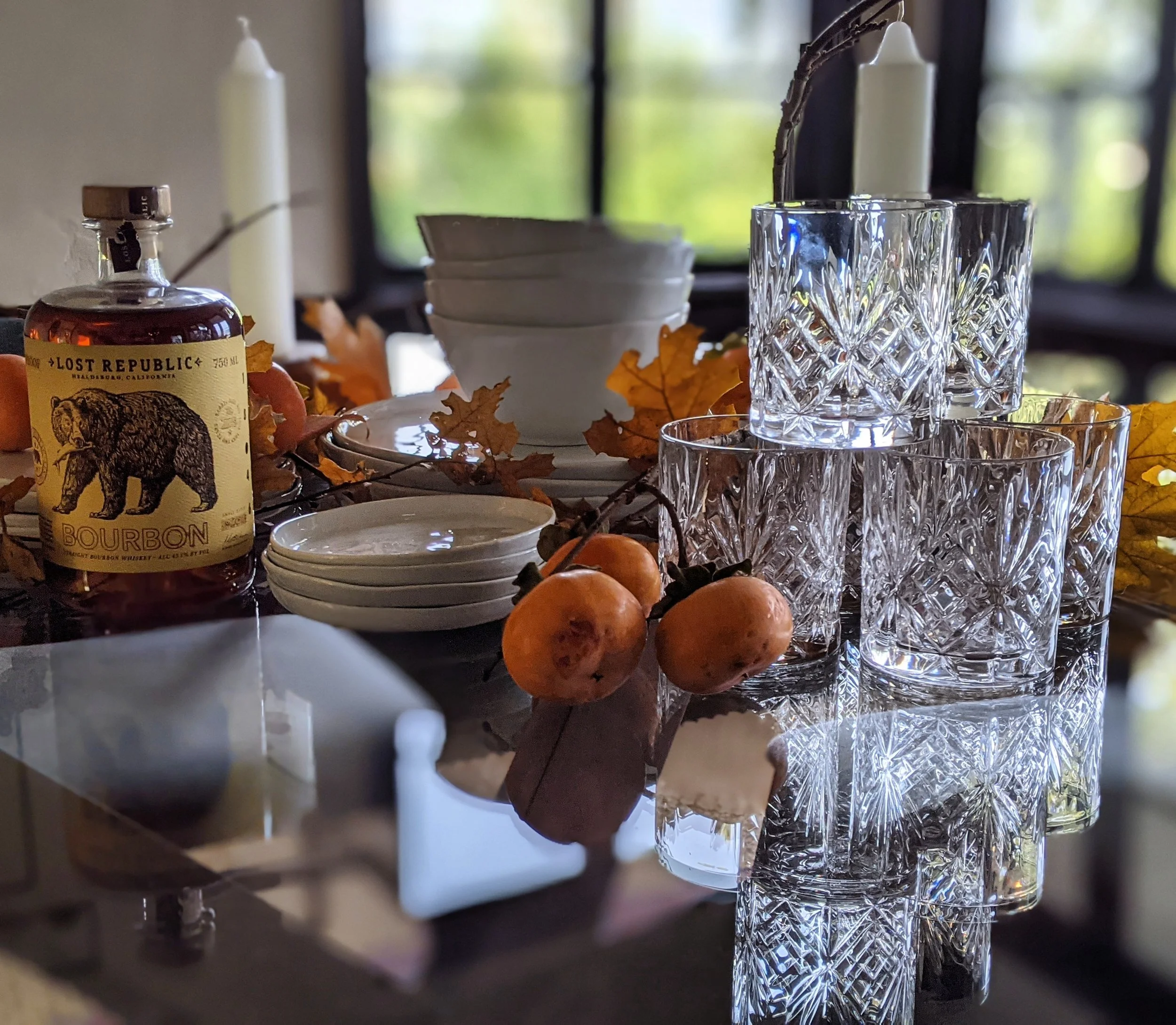 A glass bottle of Lost Republic bourbon whiskey, white bowls, orange fall leaves, dried persimmon fruits, and cut crystal glasses are arranged on a reflective surface, with a window in the background letting in natural light.