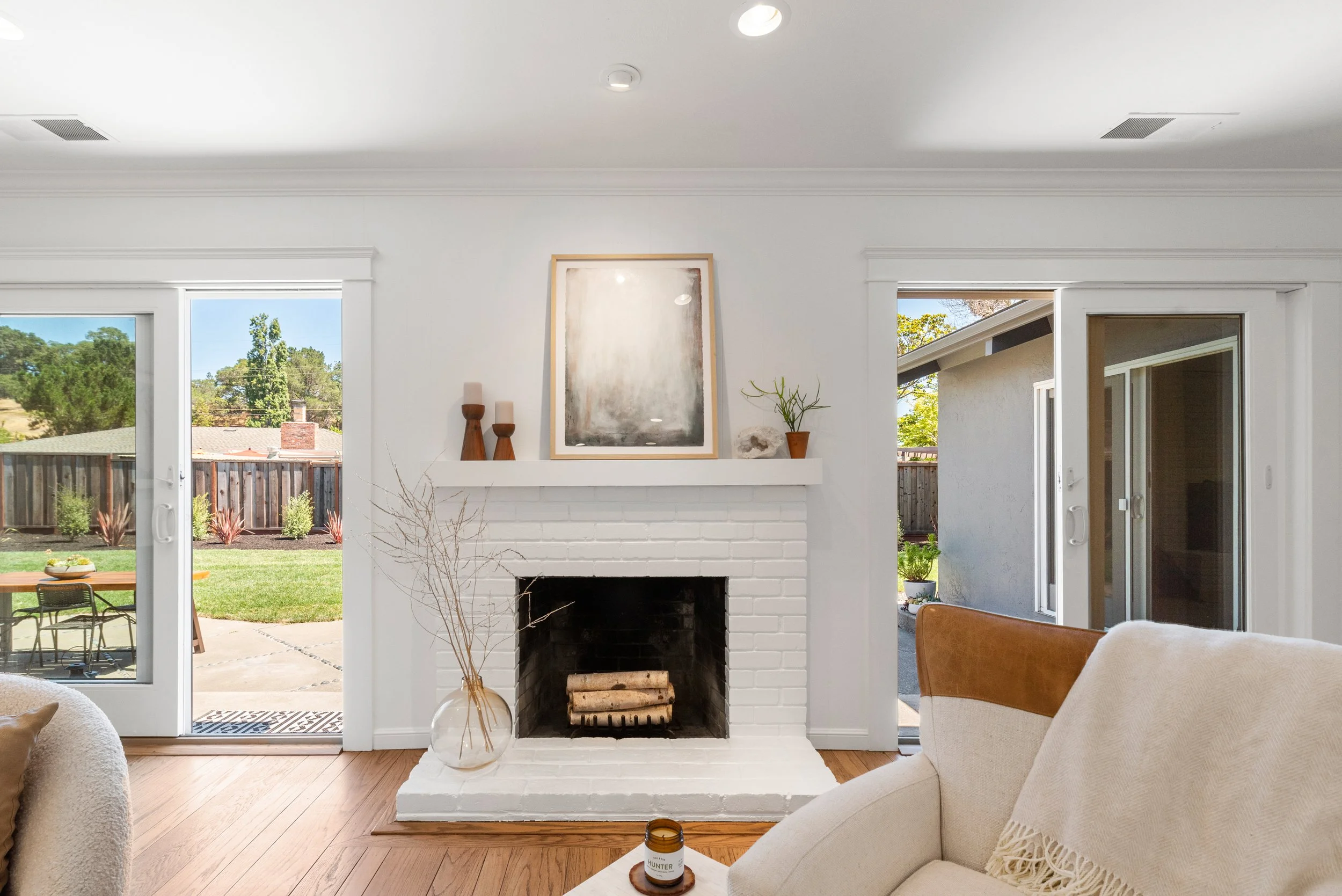 Living room with white brick fireplace, decor on mantel, glass vase with dried branches, and sliding glass doors leading to a backyard with patio and greenery.