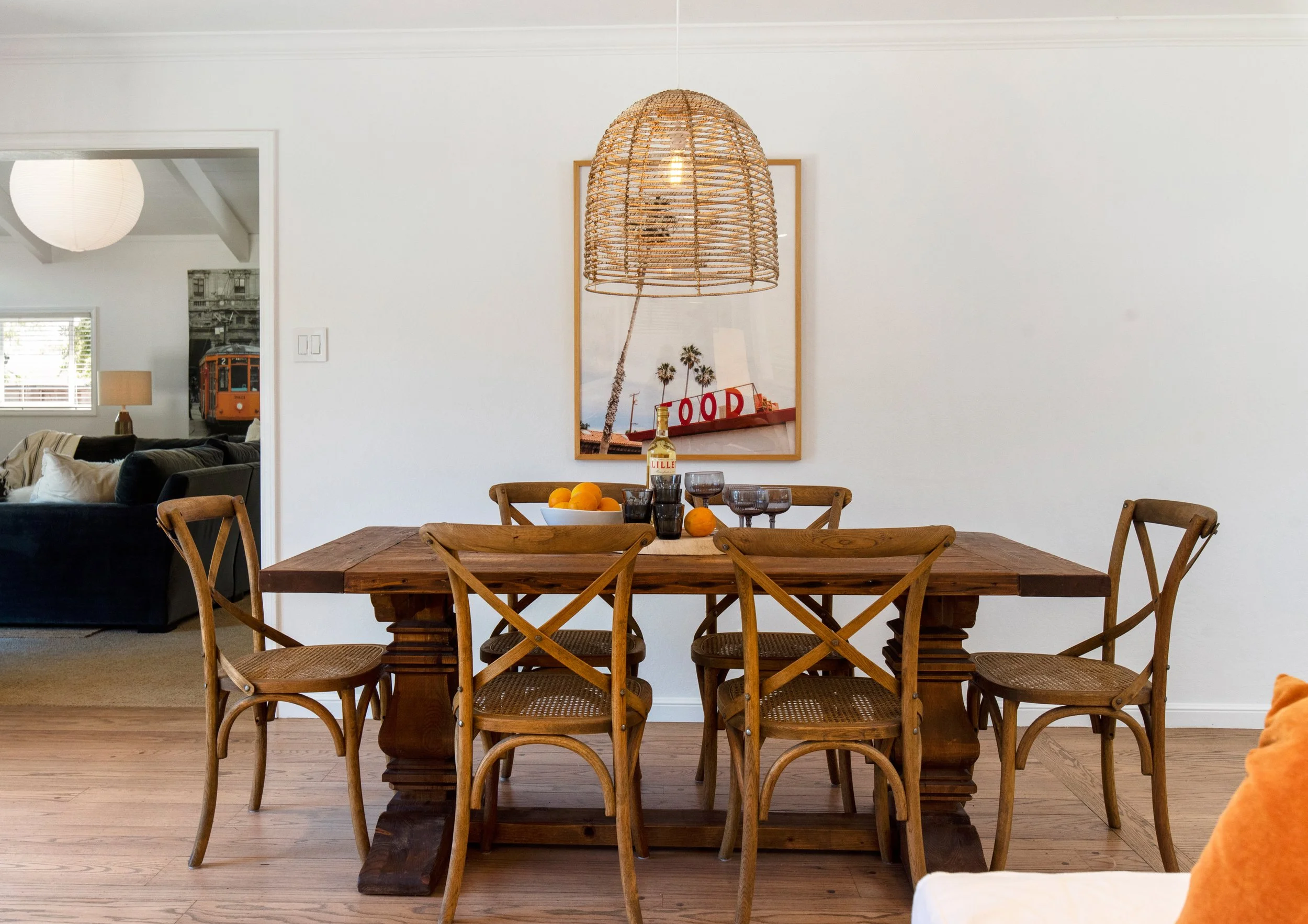 A wooden dining table with six matching chairs in a dining room. The table has a bowl of oranges, a bottle of lemonade, two glasses of dark beverage, and a few oranges on it. A large woven pendant lamp hangs above the table. On the wall behind, there is a framed picture of palm trees and the word 'FOOD' spelled out in red letters. A doorway to the left reveals a living room with a black sofa, a beige lamp, and a black-and-white photo on the wall.
