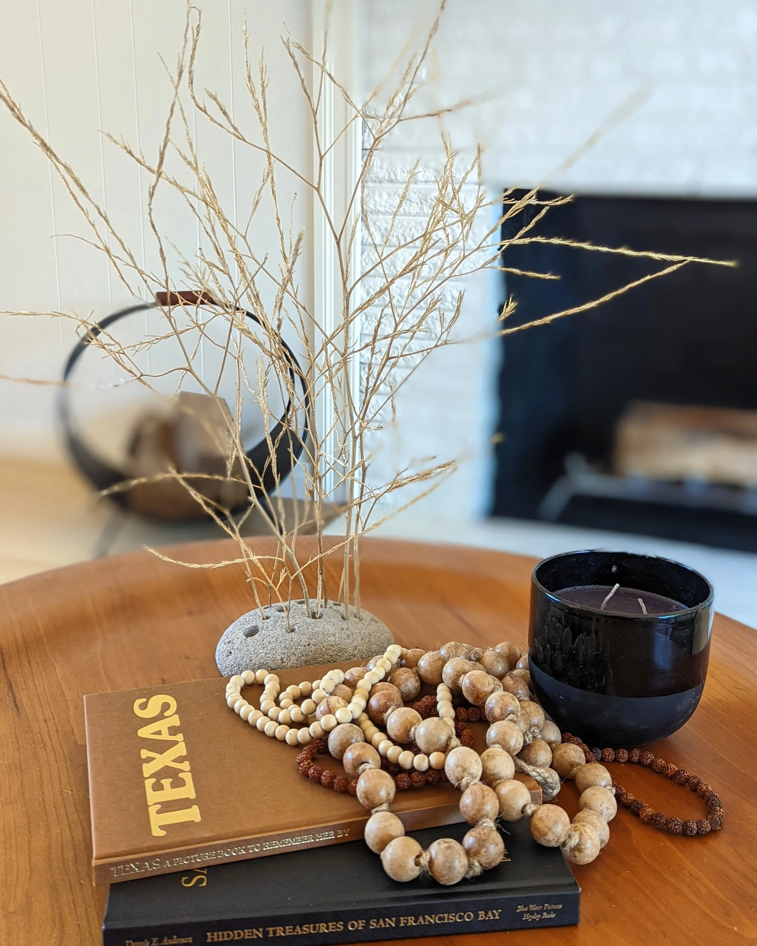 Decorative arrangement on a wooden table featuring a stack of books, a black candle holder with a candle, a string of wooden beads, a string of smaller white beads, a rock with dried grass, and a mirror in the background.