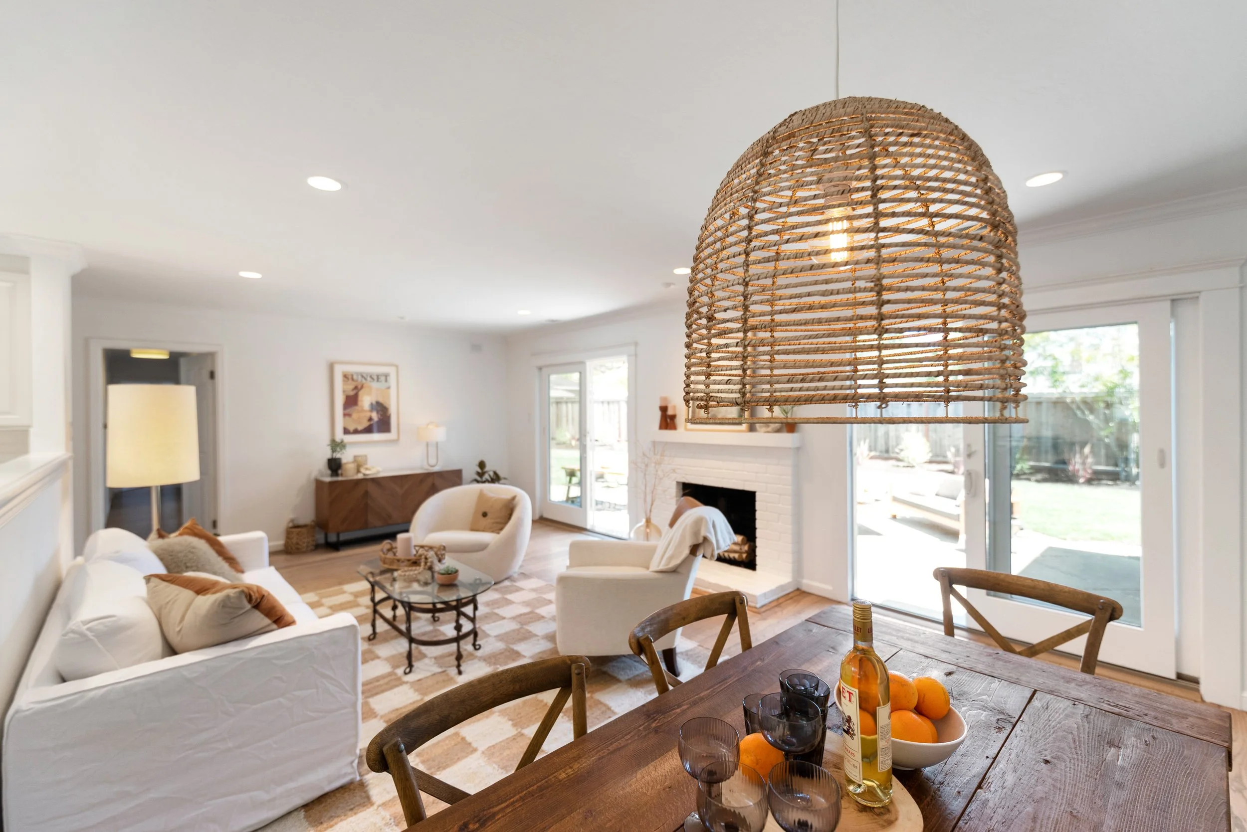 Bright living room with white walls, a fireplace, and large sliding glass doors leading to the backyard, decorated with modern and cozy furniture, a woven pendant lamp, and a wooden dining table with fruit and glasses.