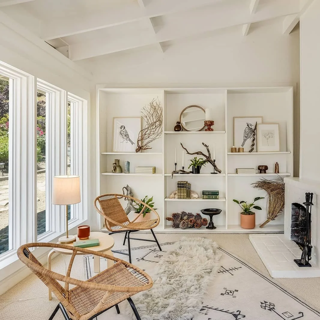 A cozy living room with large windows, a white built-in bookshelf decorated with art and plants, a fireplace, two rattan chairs, a side table with a lamp, and a cream-colored rug.