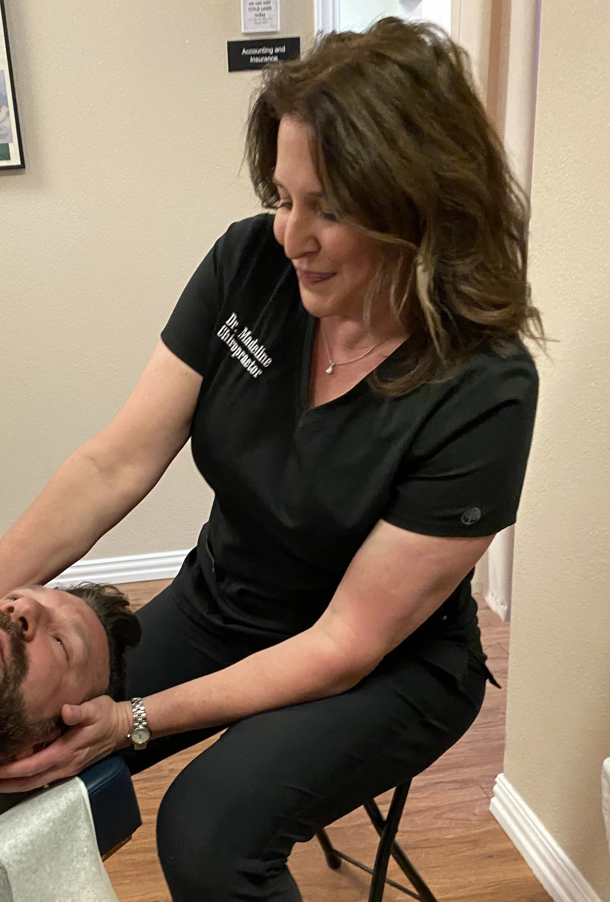 A woman giving a neck massage to a man lying on a massage table in a room with an 'Accounting and Insurance' sign on the wall.