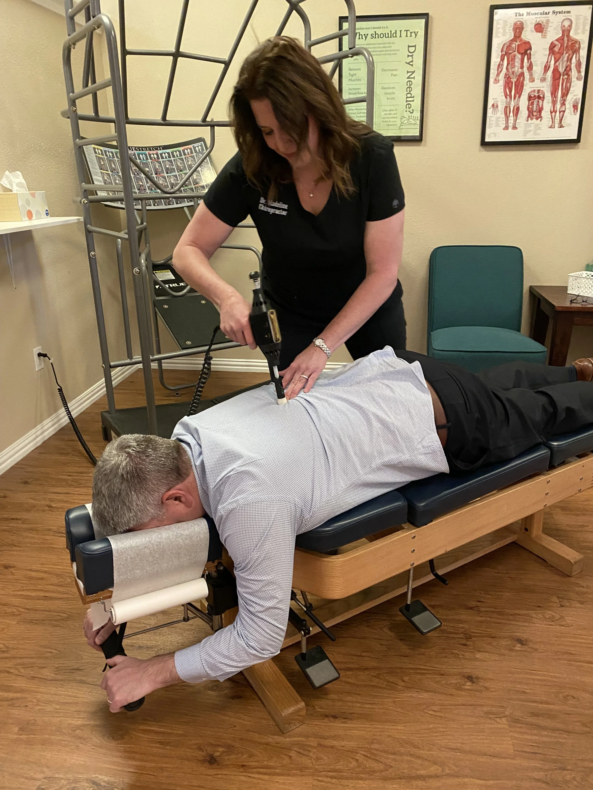 A woman performing chiropractic treatment on a man lying face down on a chiropractic table, using a chiropractic instrument in a medical office.