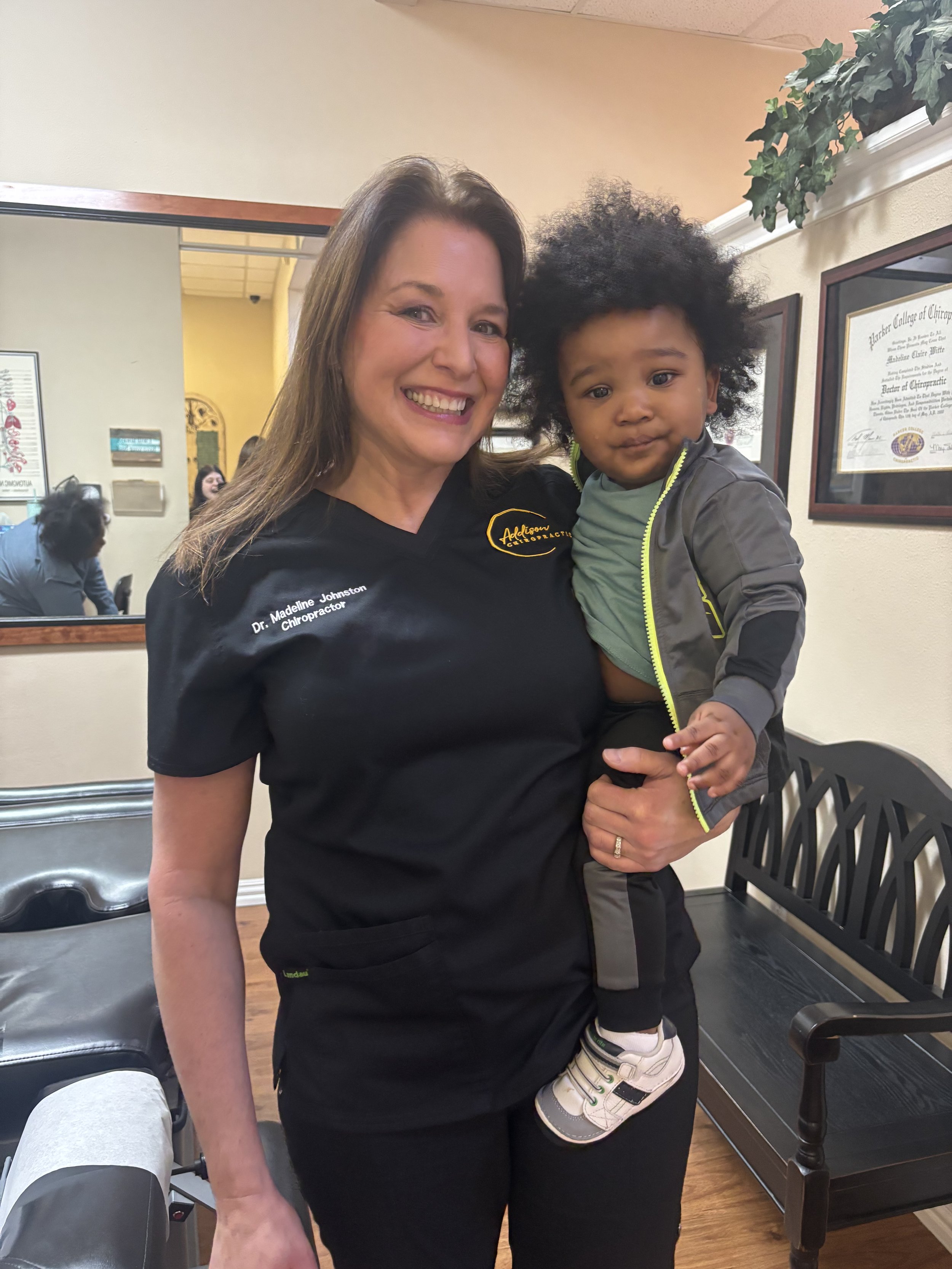 A woman with brown hair smiling while holding a young child with curly hair in a chiropractor's office.