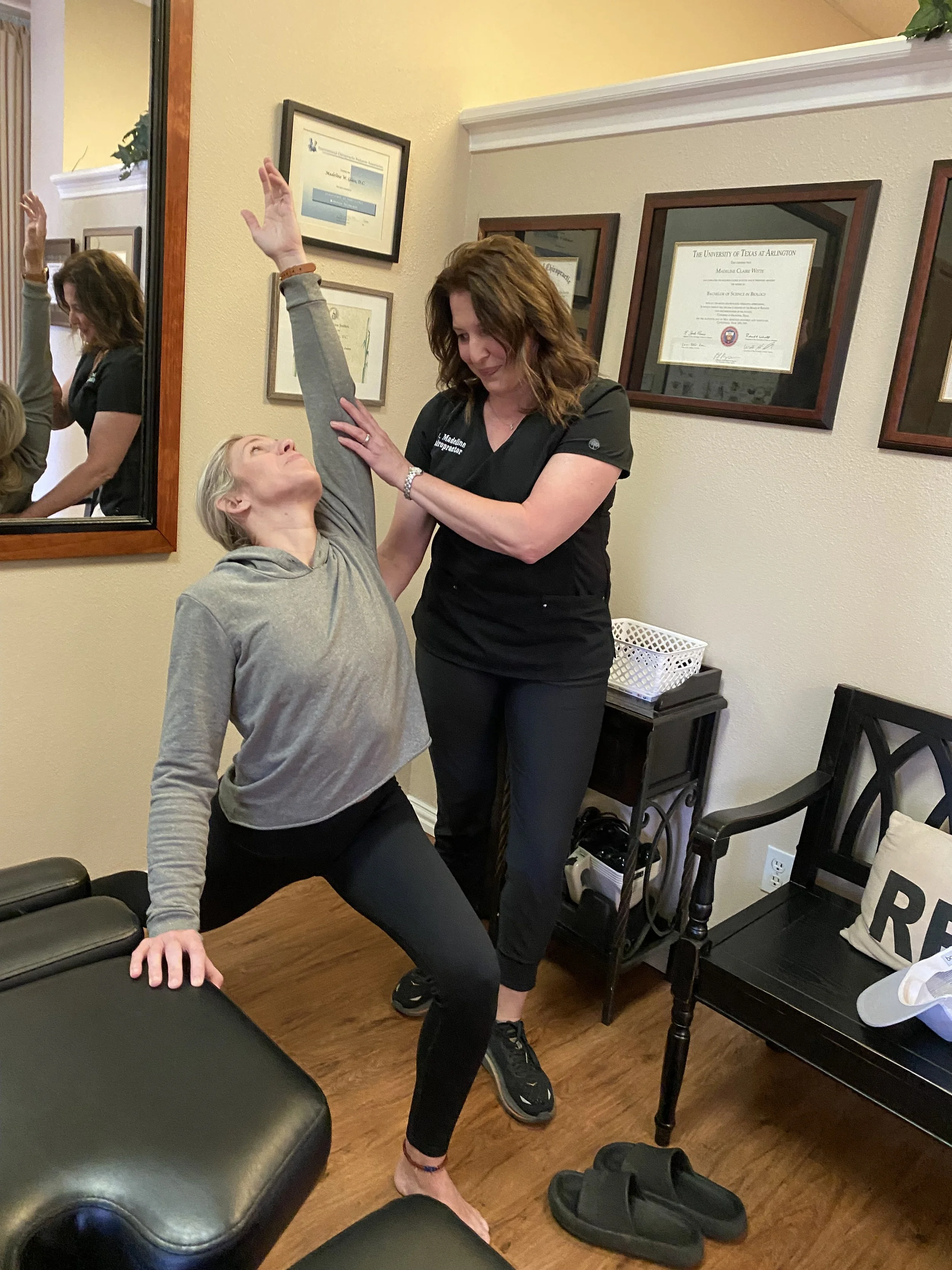 A woman practicing yoga at home while another woman, likely a healthcare provider, assists her. The healthcare provider is wearing a black shirt with writing on it. They are in a cozy room with framed diplomas on the wall, a mirror, and a wooden chair with a pillow on it.
