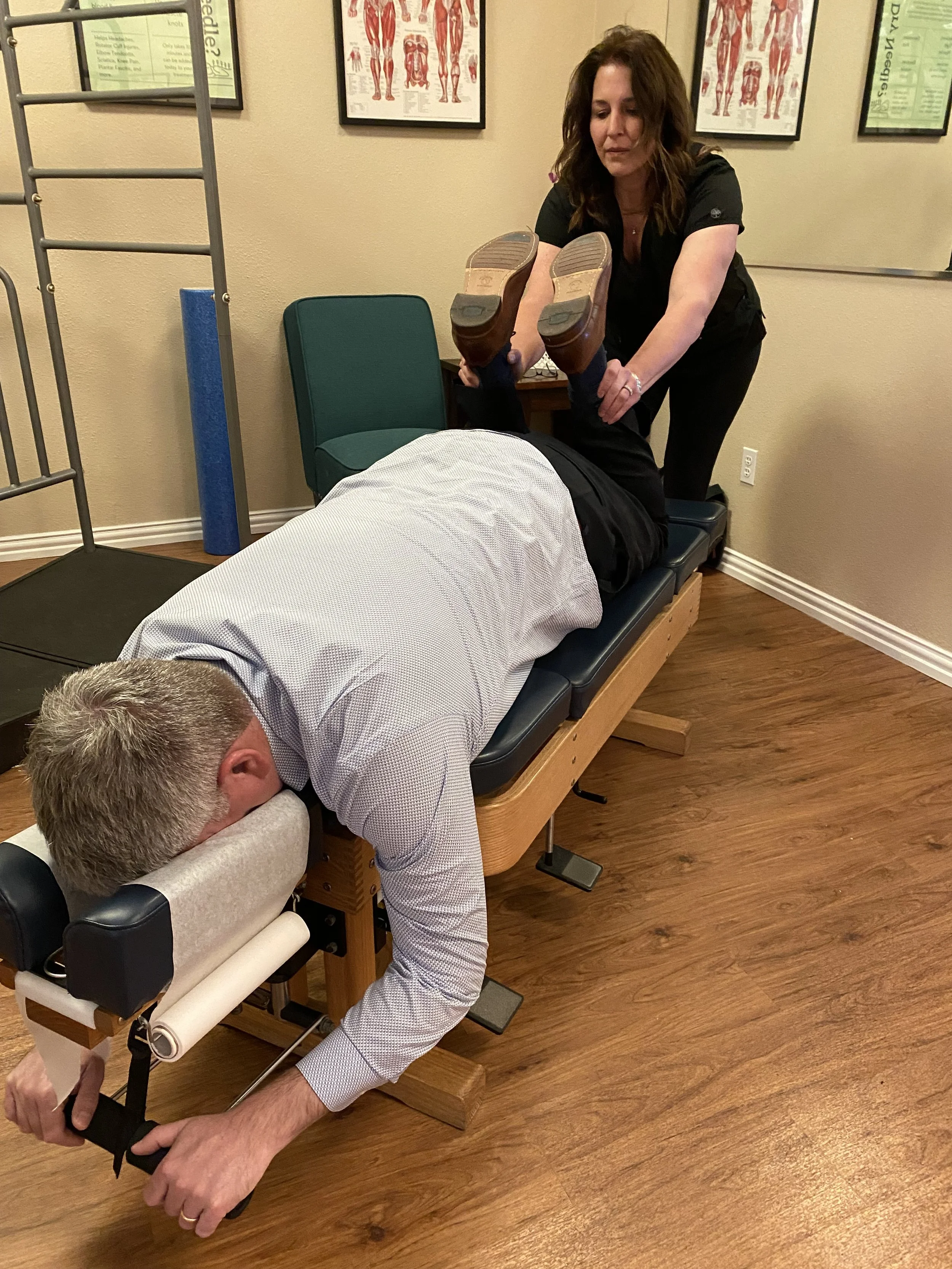 A chiropractor adjusting a patient's back on a treatment table in an office with anatomy posters on the wall.