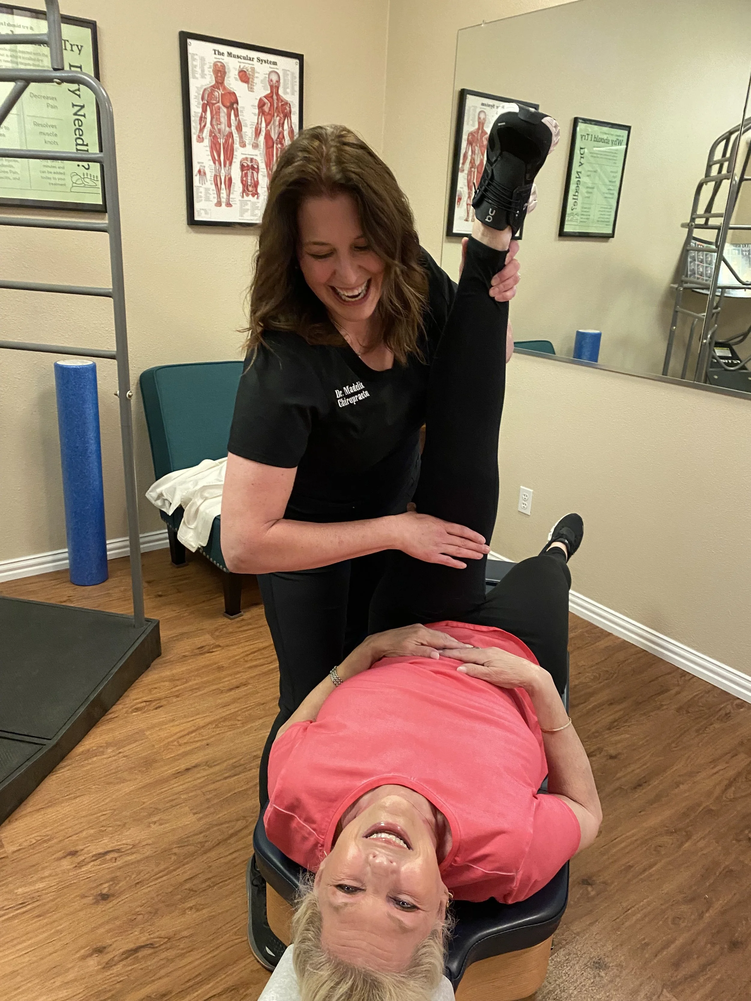 A chiropractor adjusts a woman's leg while she lies on a chiropractic table, both smiling in a clinic room with educational posters on the wall.