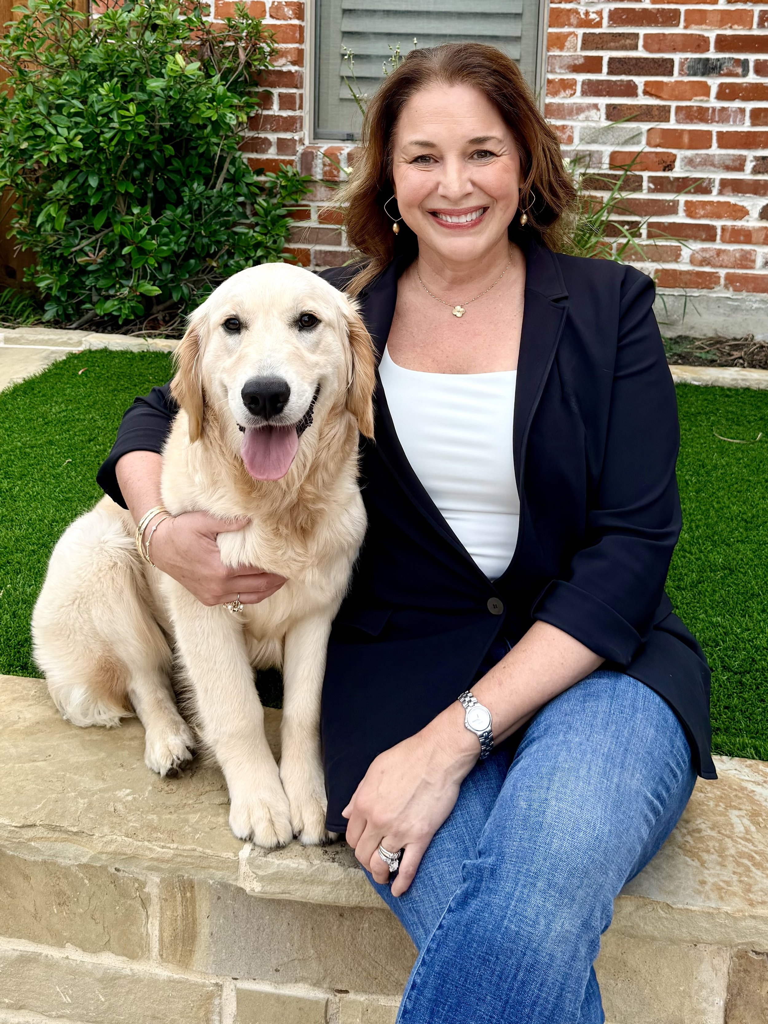 A woman smiling while sitting on a stone ledge, wearing a black blazer, white top, and blue jeans, with a watch and rings. She is holding a golden retriever puppy with light fur, sitting beside her on the ledge. The background features a brick wall, a window, and green shrubbery.