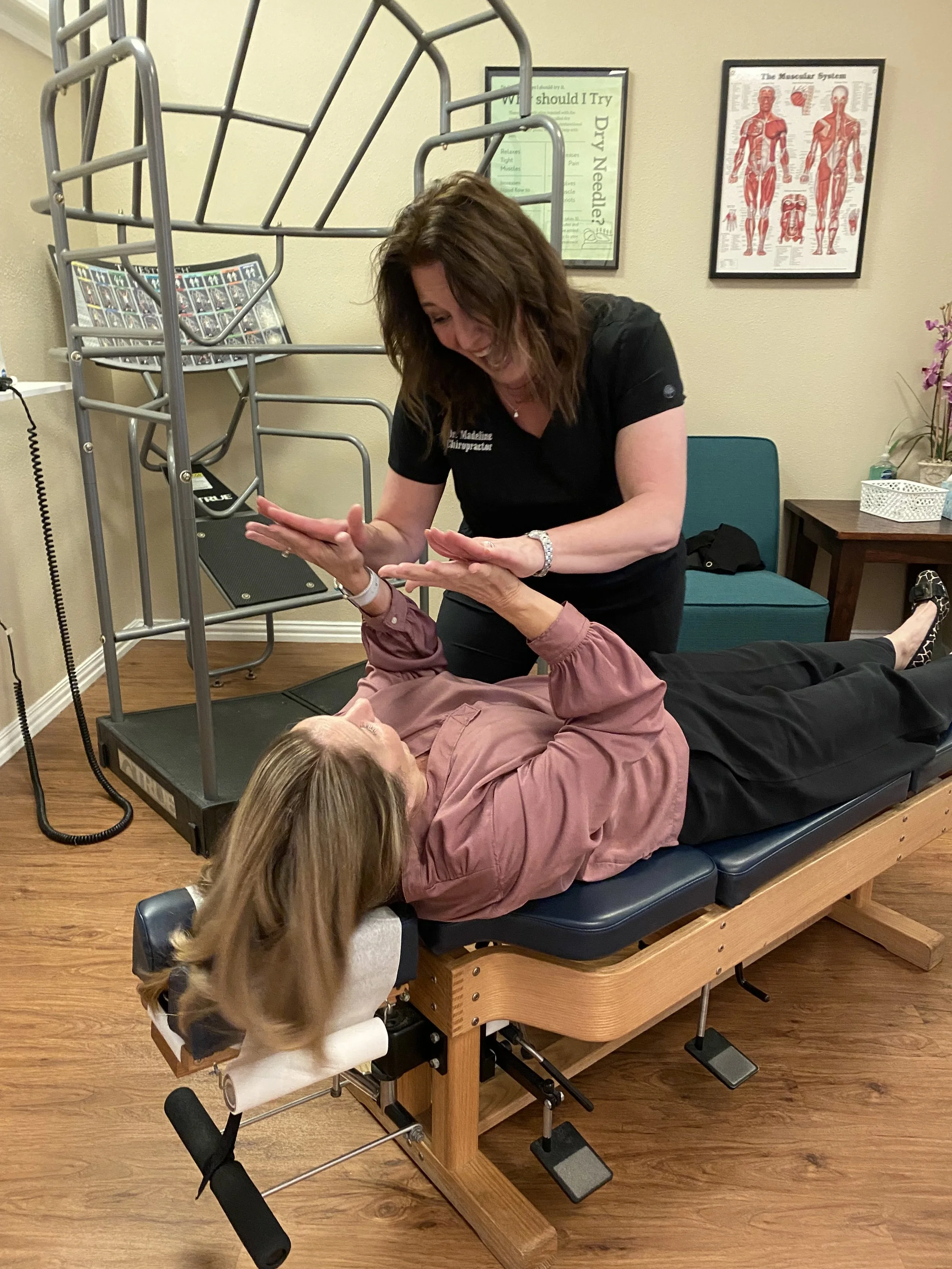 Chiropractor working with a patient lying on a chiropractic table, in an exam room with medical posters on the wall.