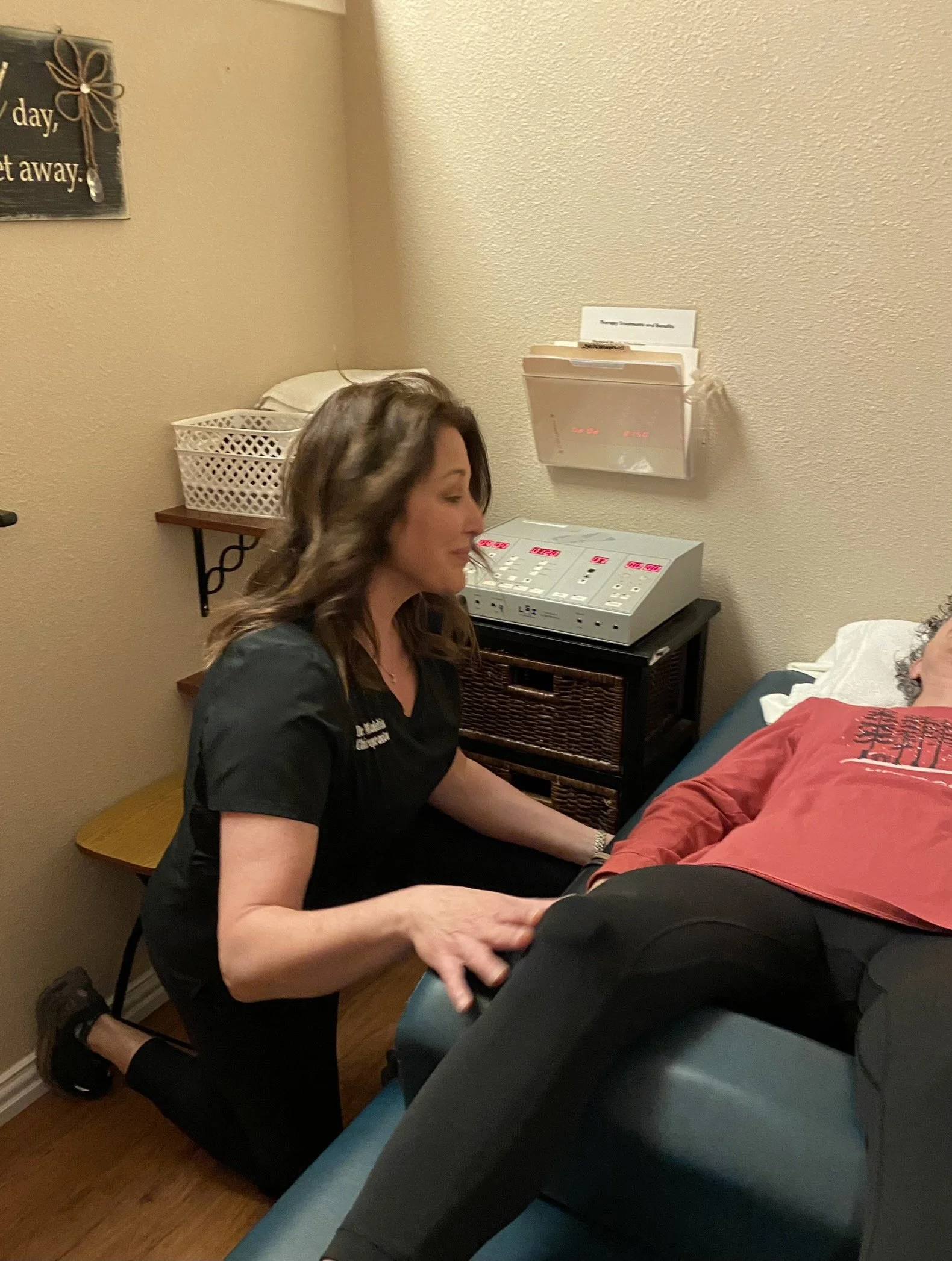 A nurse or medical professional kneeling beside a patient lying on a medical examination table, holding the patient's hand in a clinical setting.