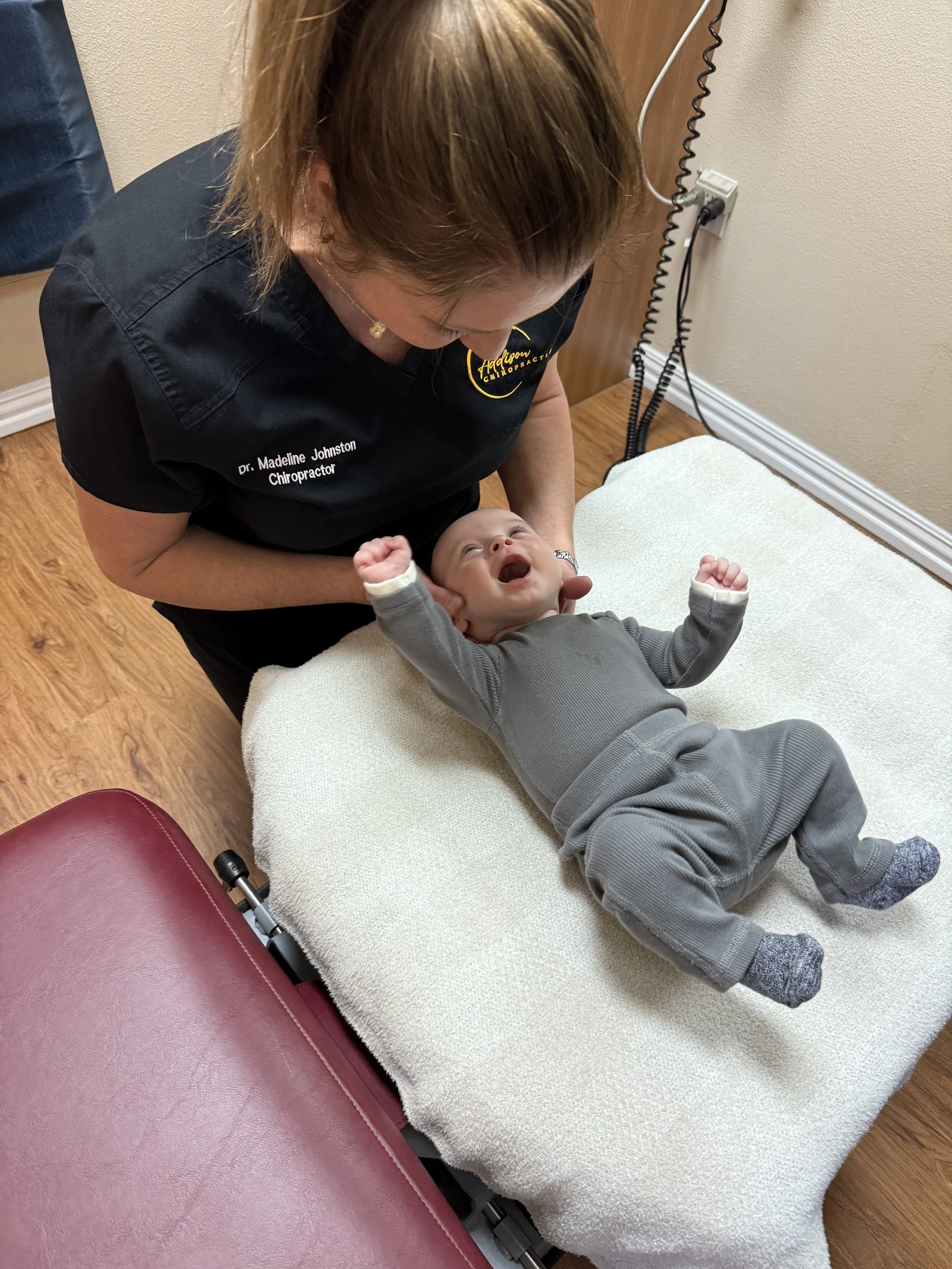 A woman with brown hair smiling while holding a young child with curly hair in a chiropractor's office.