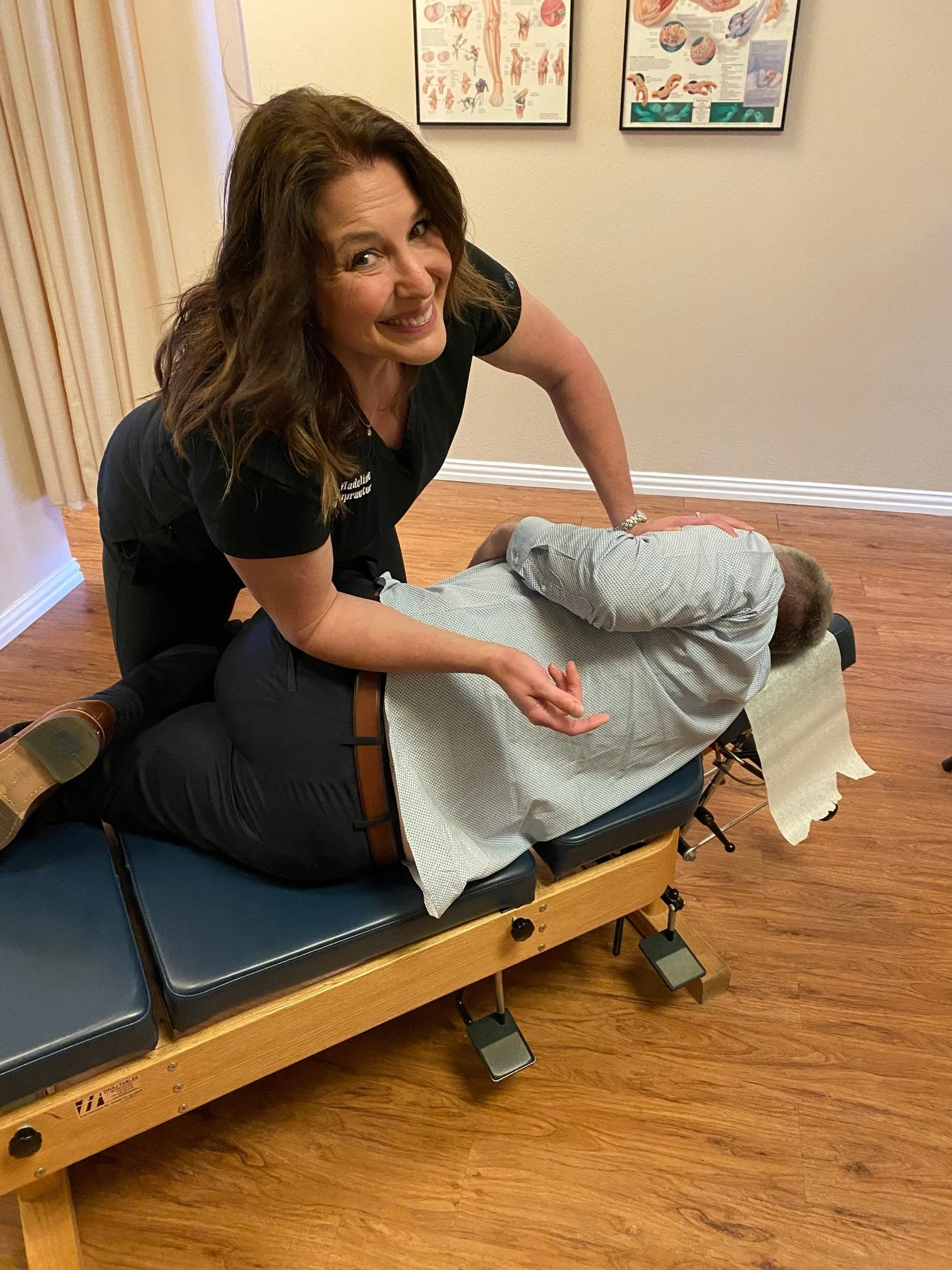 A chiropractor performing an adjustment on a patient lying face down on a chiropractic table in an office with anatomical posters on the wall.