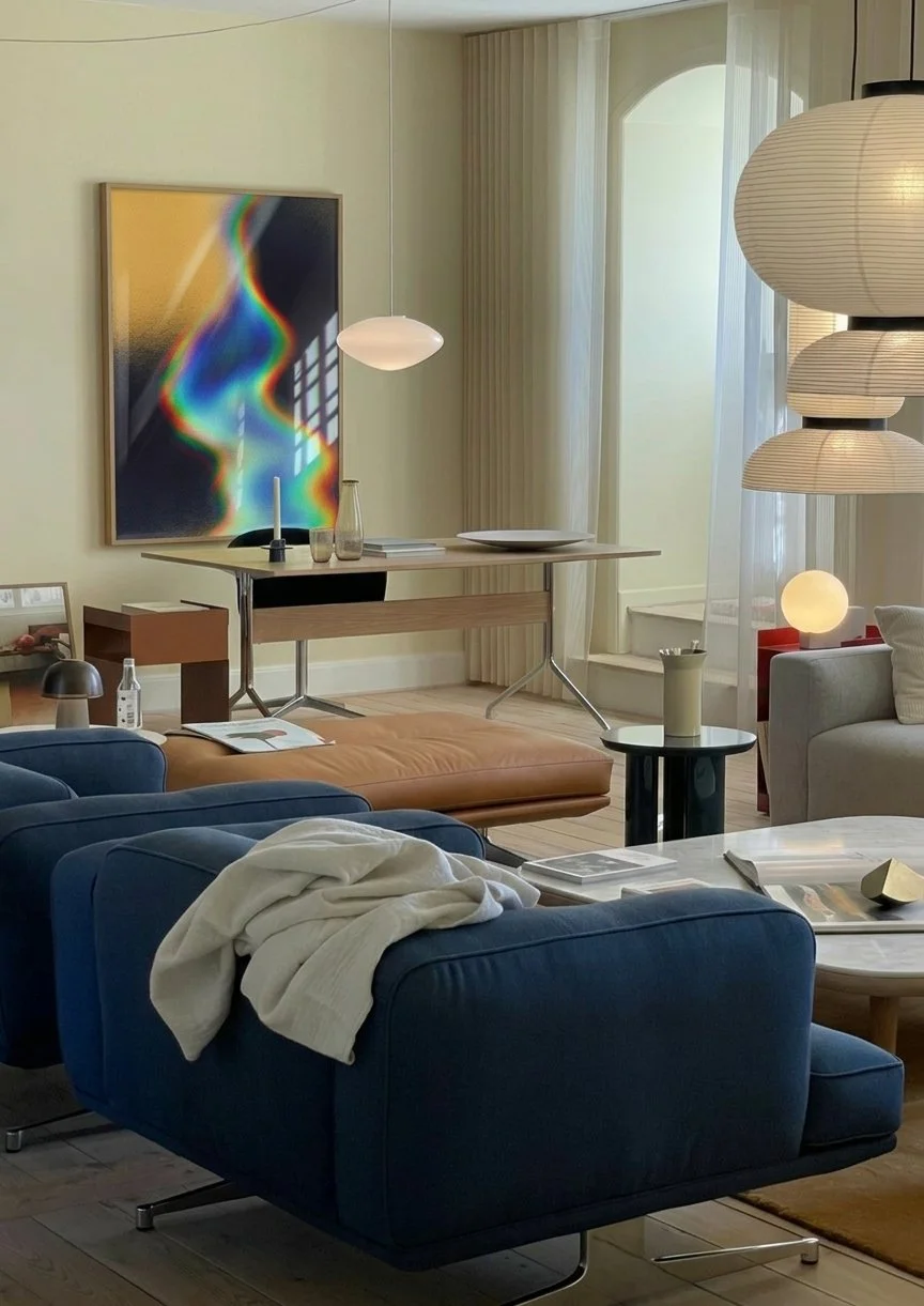Living room with blue sofas, a tan ottoman, a small black side table with a beige vase, a white marble coffee table with magazines, a beige armchair, paper lanterns, wall art, and sheer curtains.