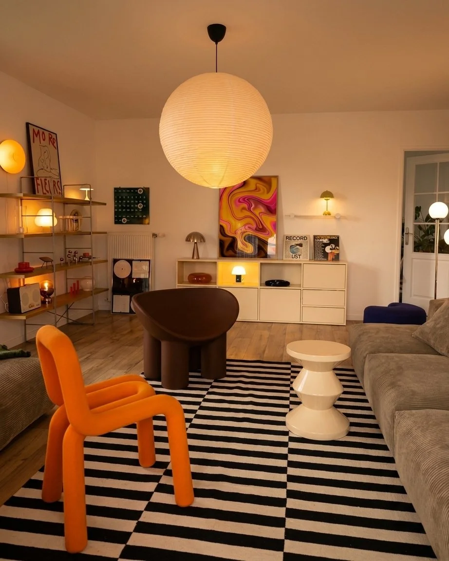 A living room with a large paper lantern ceiling light, a black armchair, an orange children's chair, a gray sofa, a white side table, and a striped black-and-white rug. The room has modern decor, wall art, shelves with lamps, and a vinyl record play