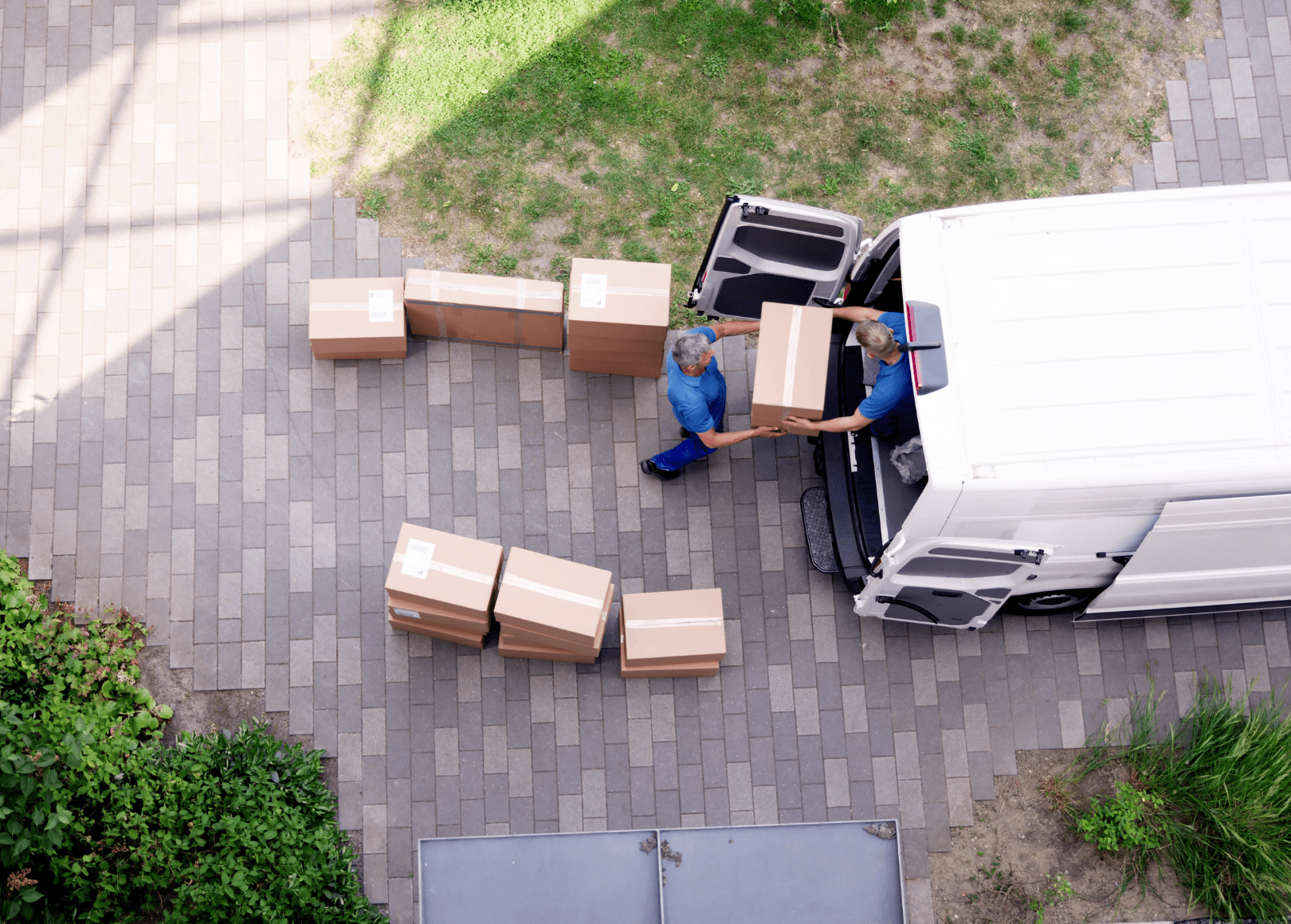 Two movers in blue uniforms unloading boxes from a white moving van onto a brick driveway, with some boxes placed on the ground and others being handed over.