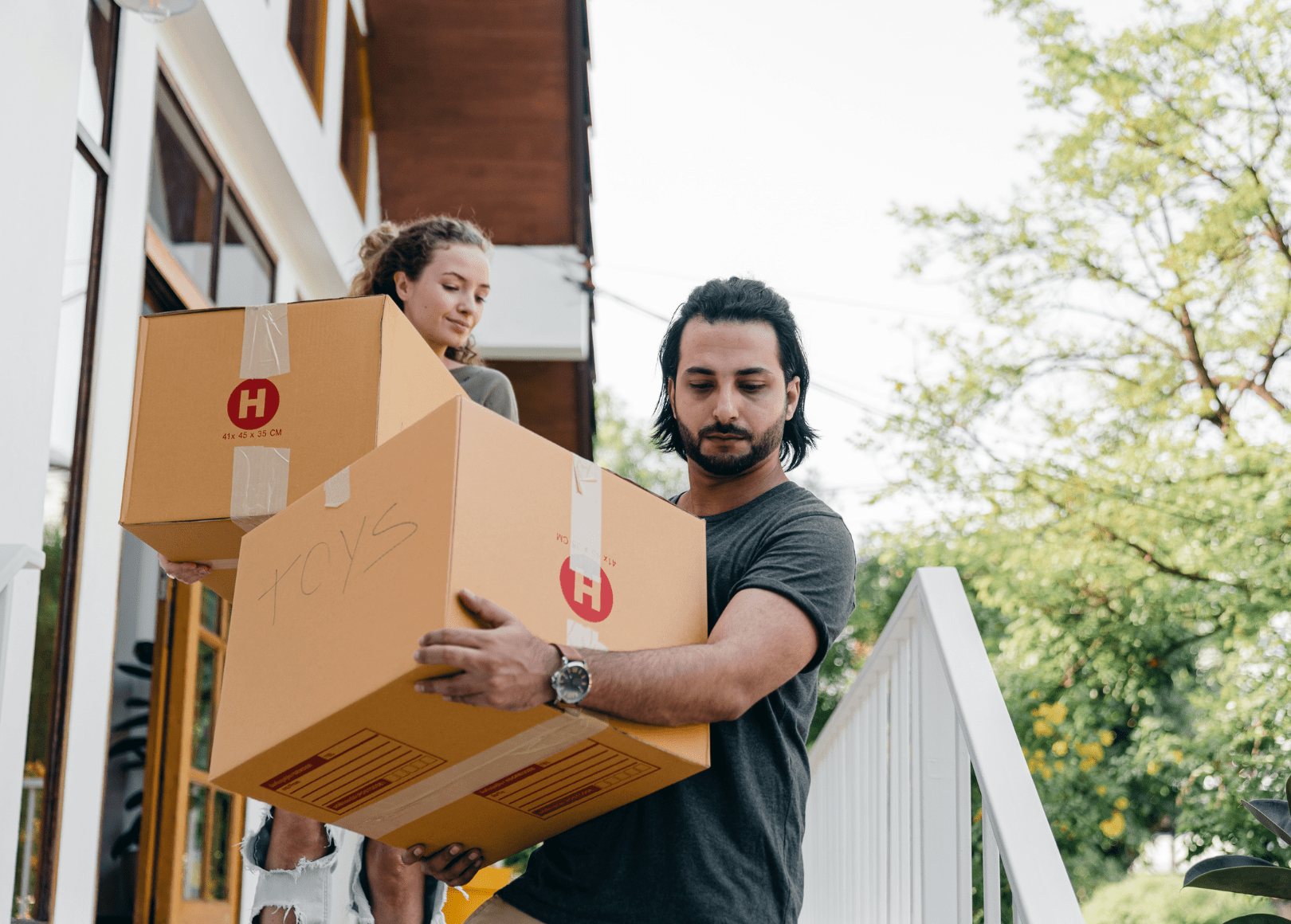 A man and a woman moving boxes outside a house, the man is holding a large cardboard box, and the woman is carrying a smaller box, both are on the porch with green trees in the background.