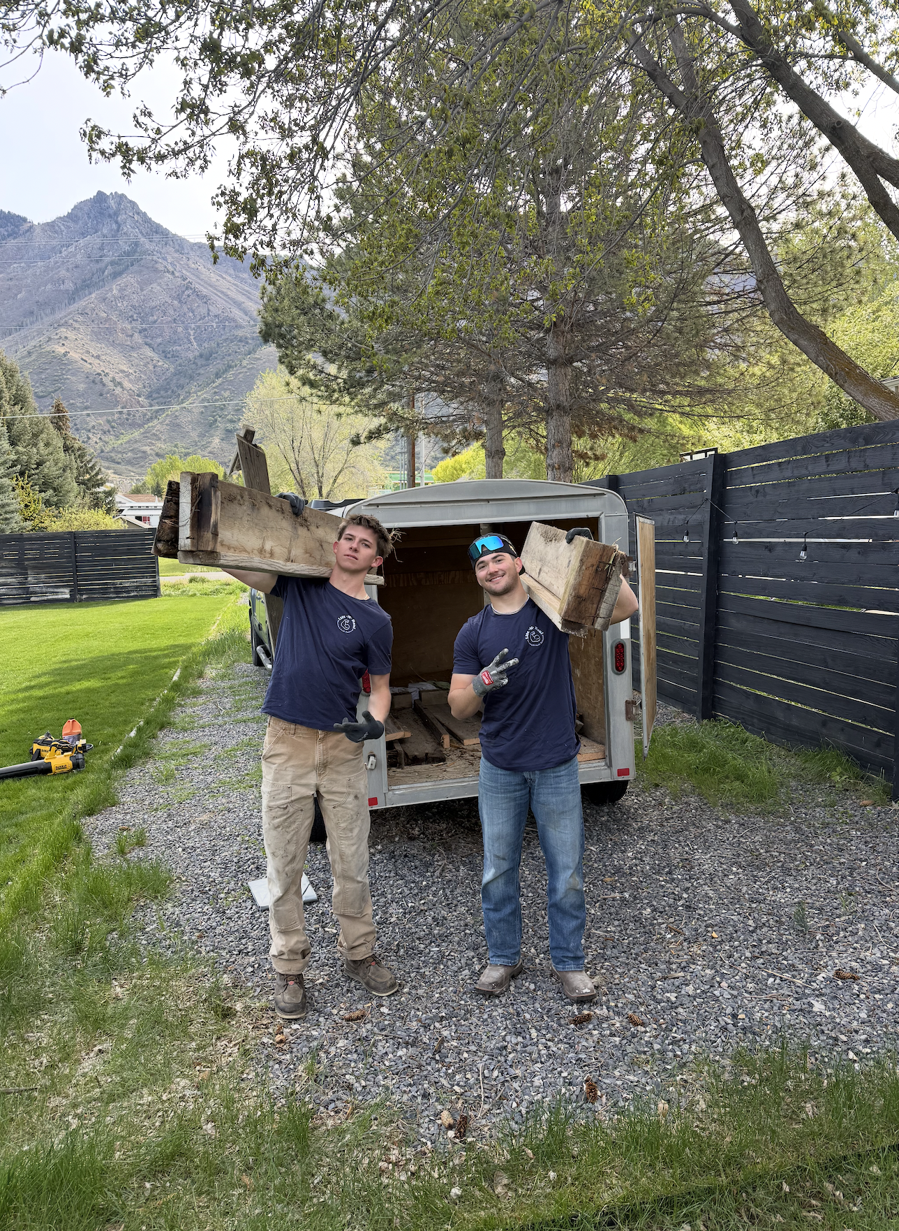 Two men are moving furniture in a garage. One is carrying a white cabinet, and the other is standing with his back to the camera. The garage contains moving boxes, a wooden panel, and packaging materials.