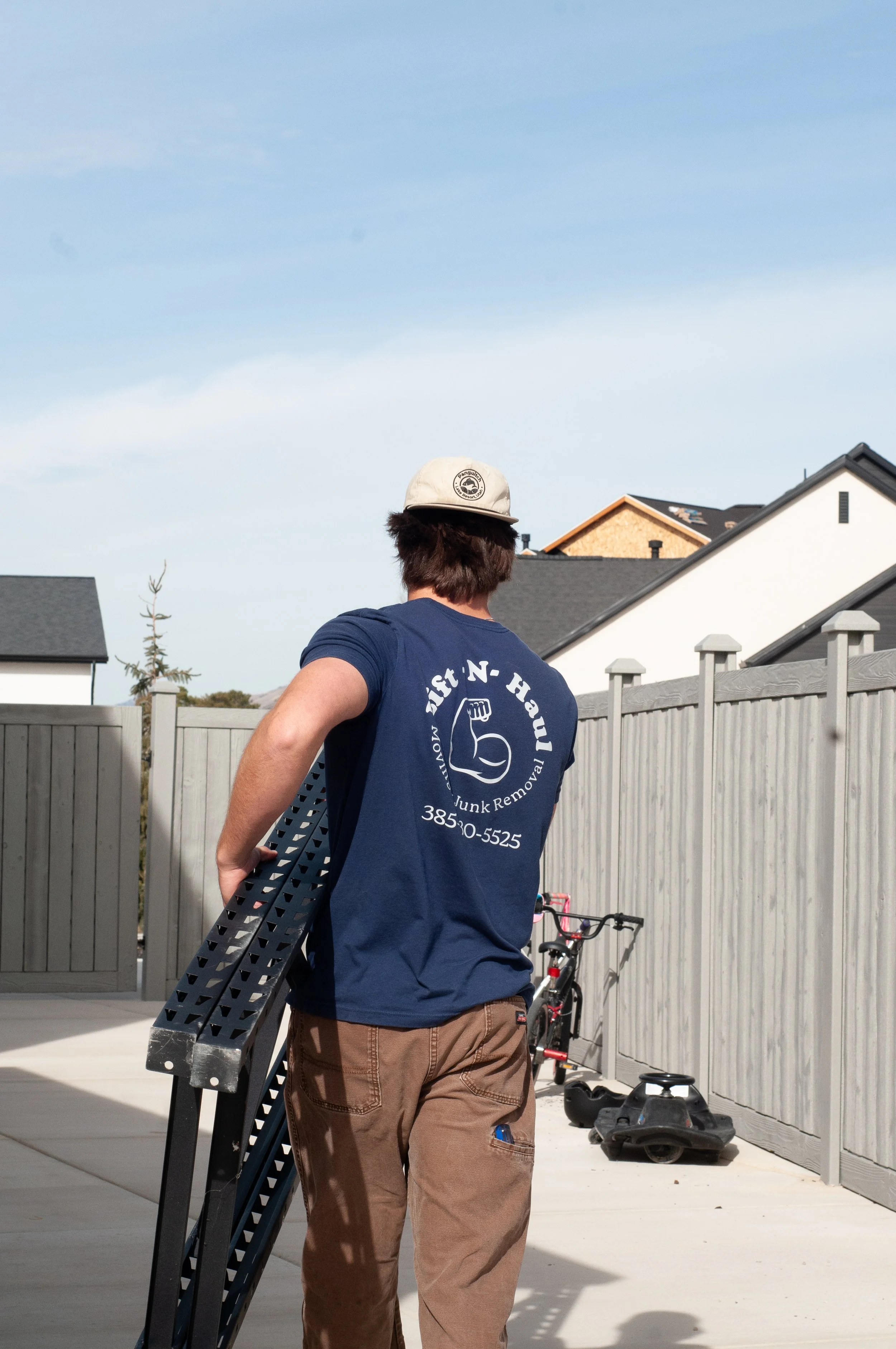A man and a woman moving boxes outside a house, the man is holding a large cardboard box, and the woman is carrying a smaller box, both are on the porch with green trees in the background.