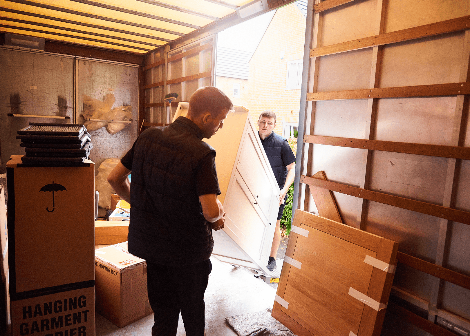 Two men are moving furniture in a garage. One is carrying a white cabinet, and the other is standing with his back to the camera. The garage contains moving boxes, a wooden panel, and packaging materials.