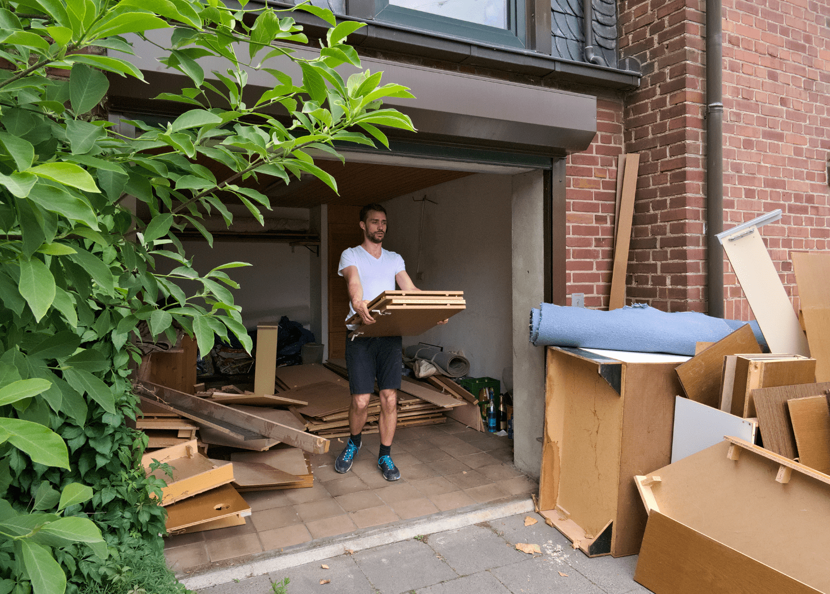 A man is moving boxes and furniture out of a garage, with scattered items and furniture pieces around him.