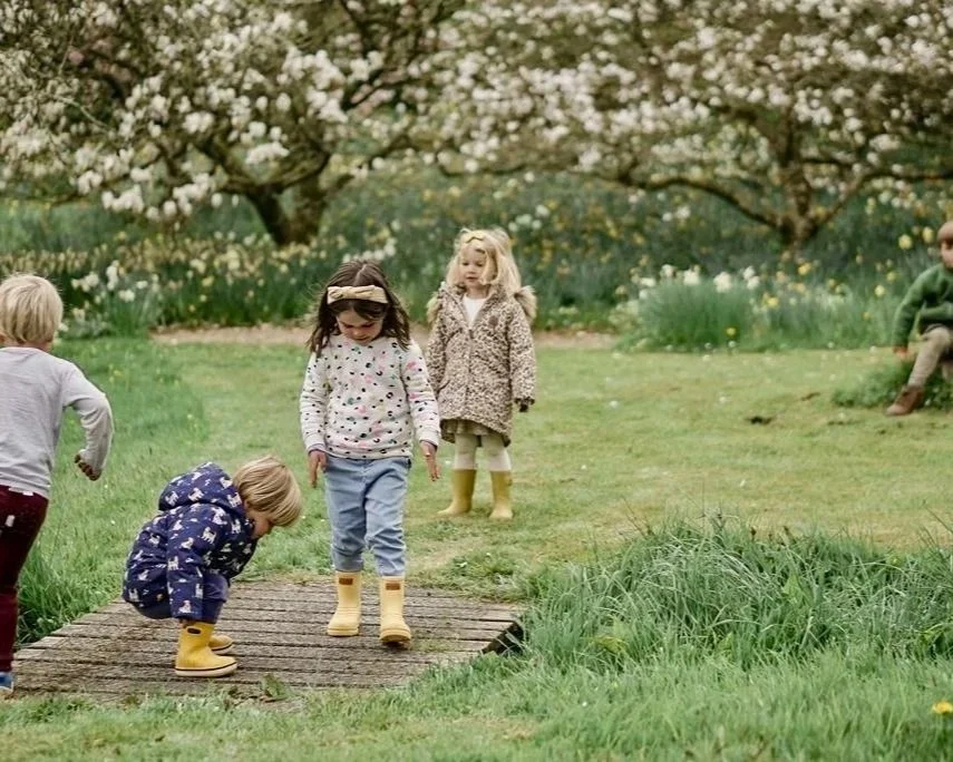 Children playing outdoors on a grassy field with blooming trees in the background, some wearing yellow rain boots.