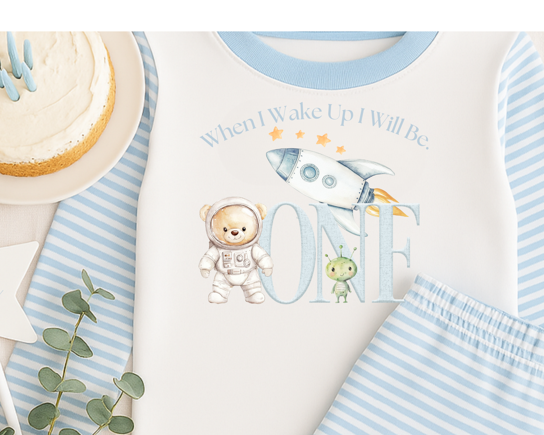 A baby onesie with space-themed illustrations including a rocket, a bear dressed as an astronaut, a cute alien, and text that reads "When I Wake Up I Will Be ONE." The onesie is light-colored with blue accents around the collar, and it is displayed with a blue and white striped cardigan and a plate with a slice of cake garnished with blue icing.