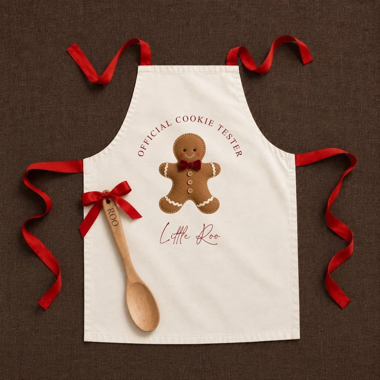 White apron with red ribbons and a gingerbread man design, labeled "Official Cookie Tester" and "Little Roo." A wooden spoon tied with a red ribbon and a small wooden tag that says "Roo" are also on the apron.