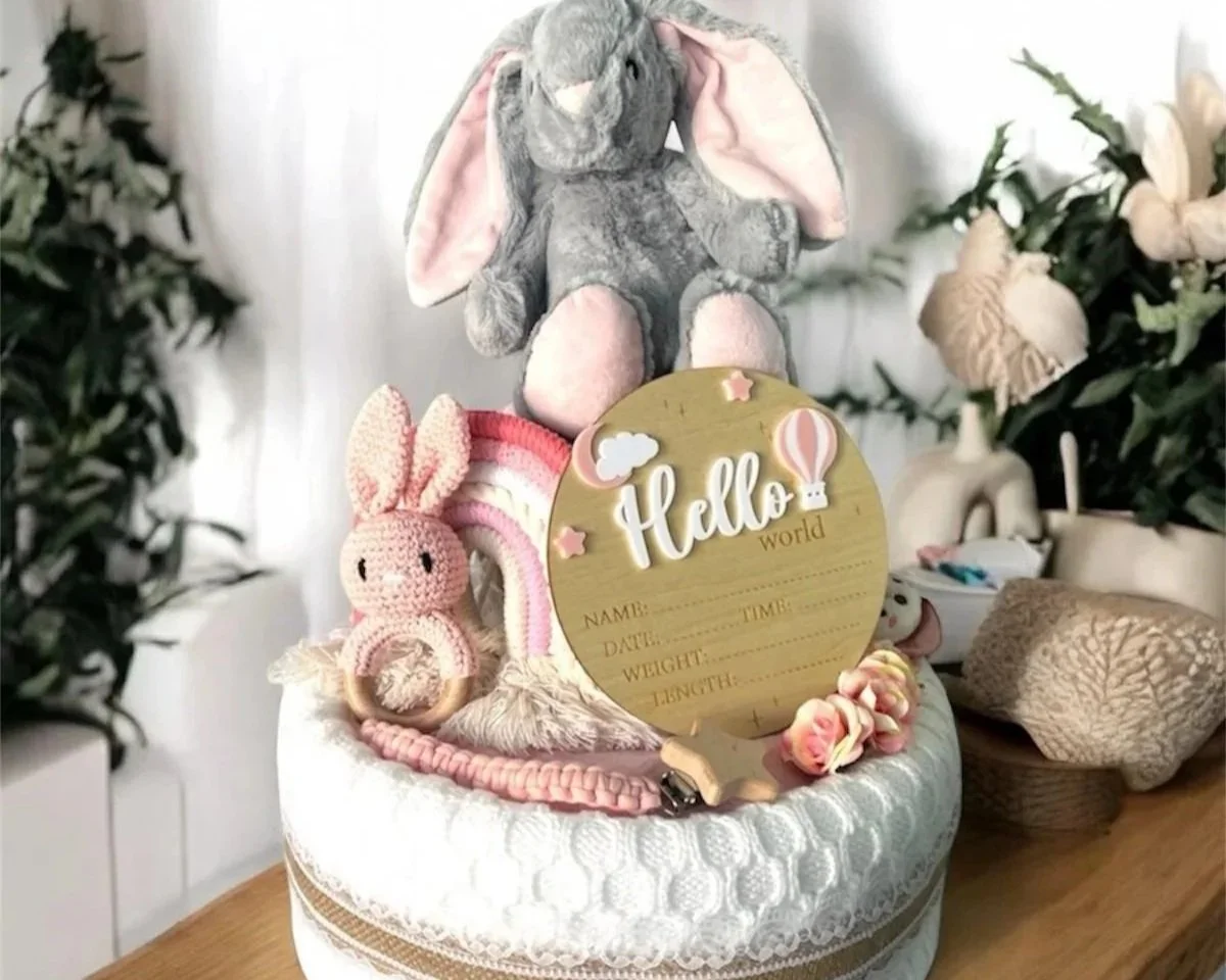 Decorative baby nursery items including a plush gray elephant with big ears, a pink knitted bunny rattle, a round wooden sign with 'Hello world' text, and a pink and white rainbow, all arranged on a white textured round surface with soft surrounding decor.