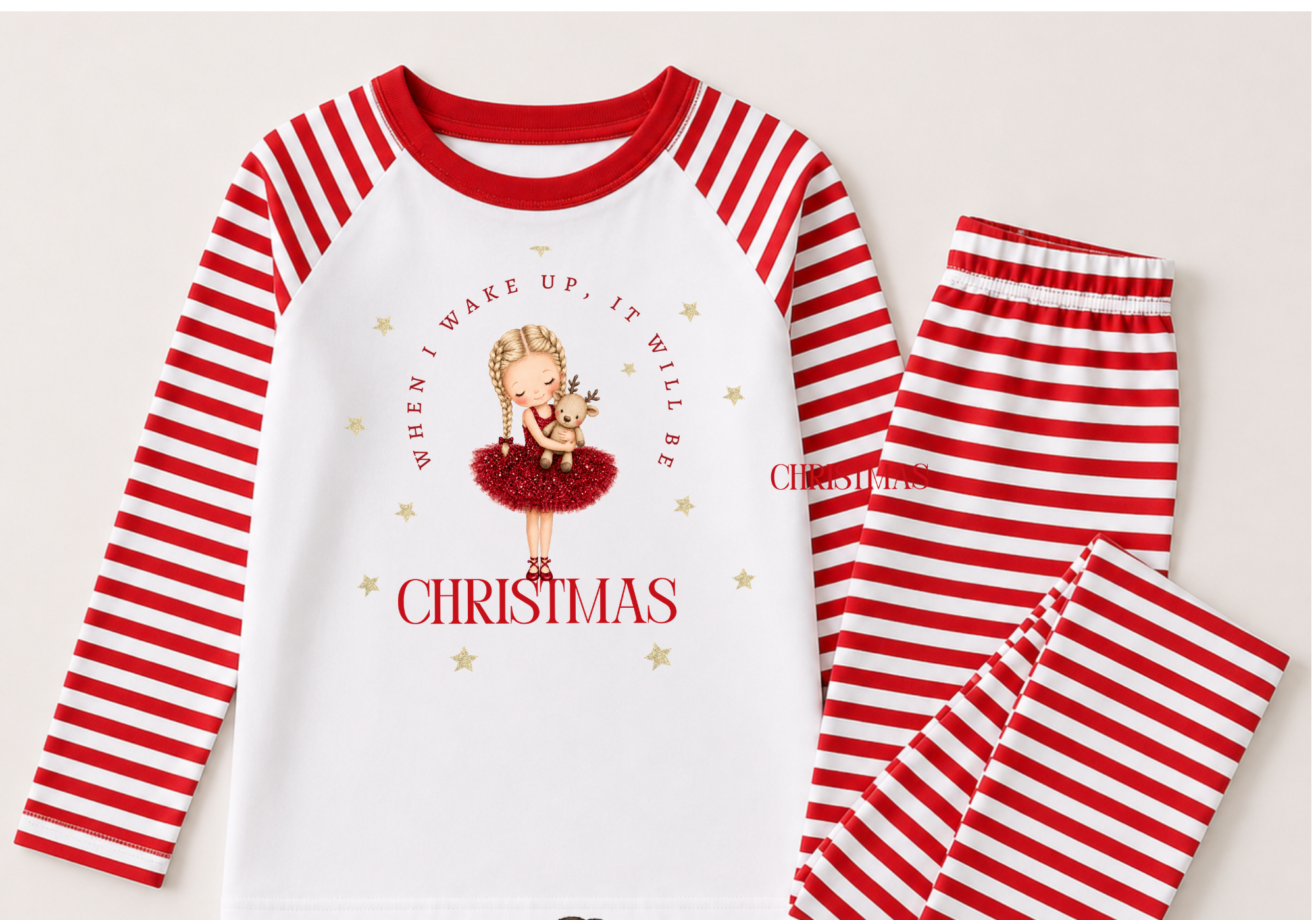 Red and white striped pajamas with a Christmas-themed graphic on the front, featuring a girl with braids holding a reindeer, surrounded by gold stars. The shirt has the text "When I wake up, it will be Christmas." The pajama pants have red and white horizontal stripes.