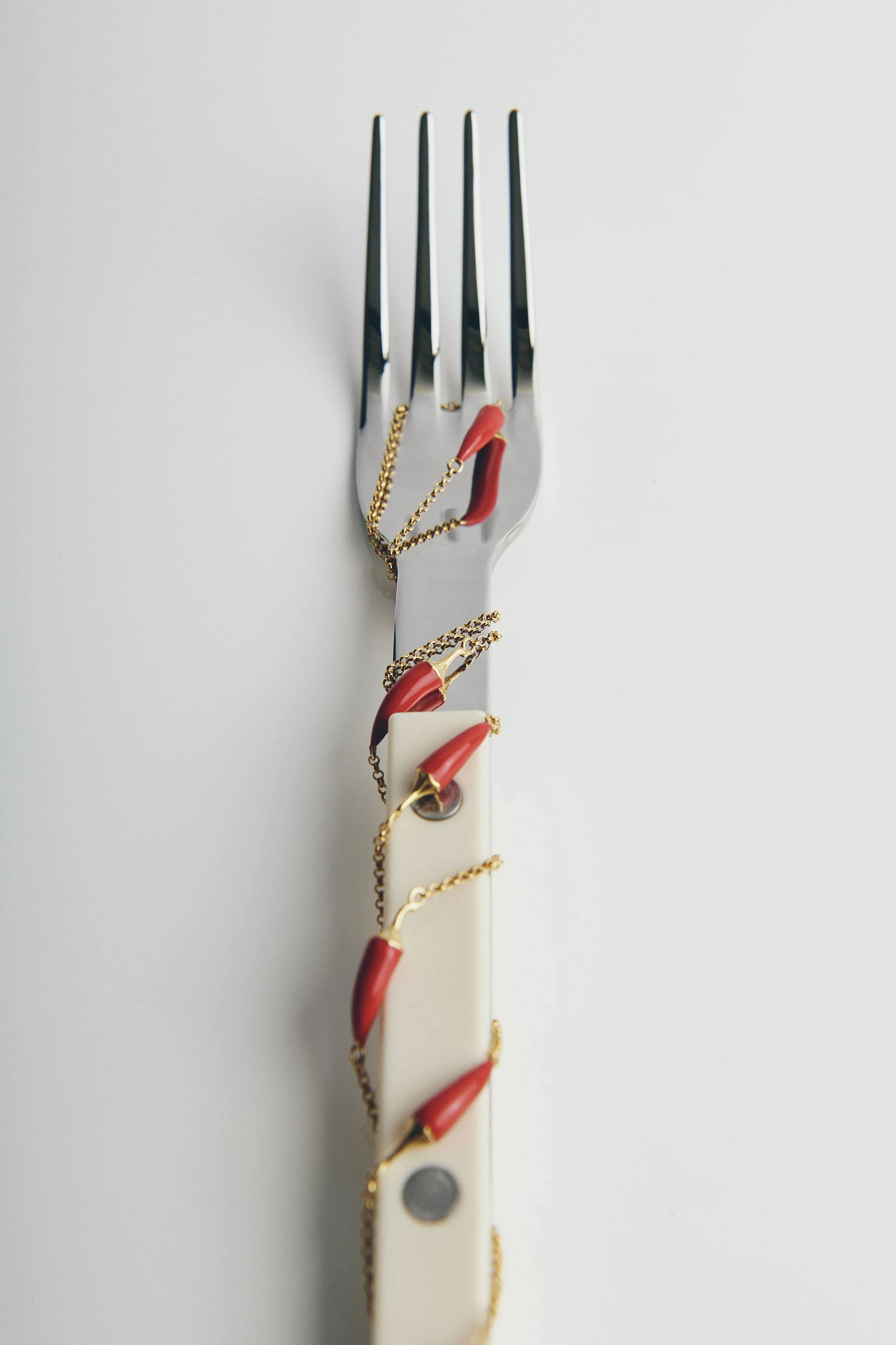 A metal fork with a gold chain and red chili pepper charms wrapped around it, with a white handle also decorated with the same charms, on a plain white background.