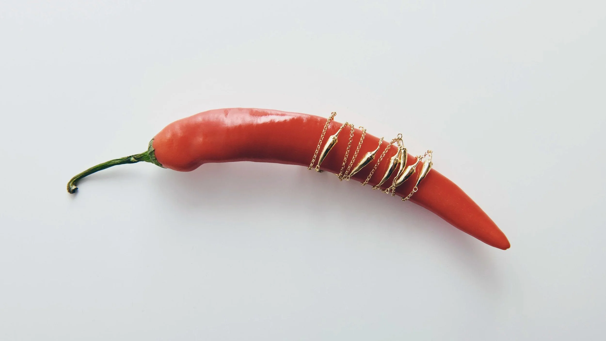 A red chili pepper with the stem on the left, wrapped with various gold jewelry chains and rings.
