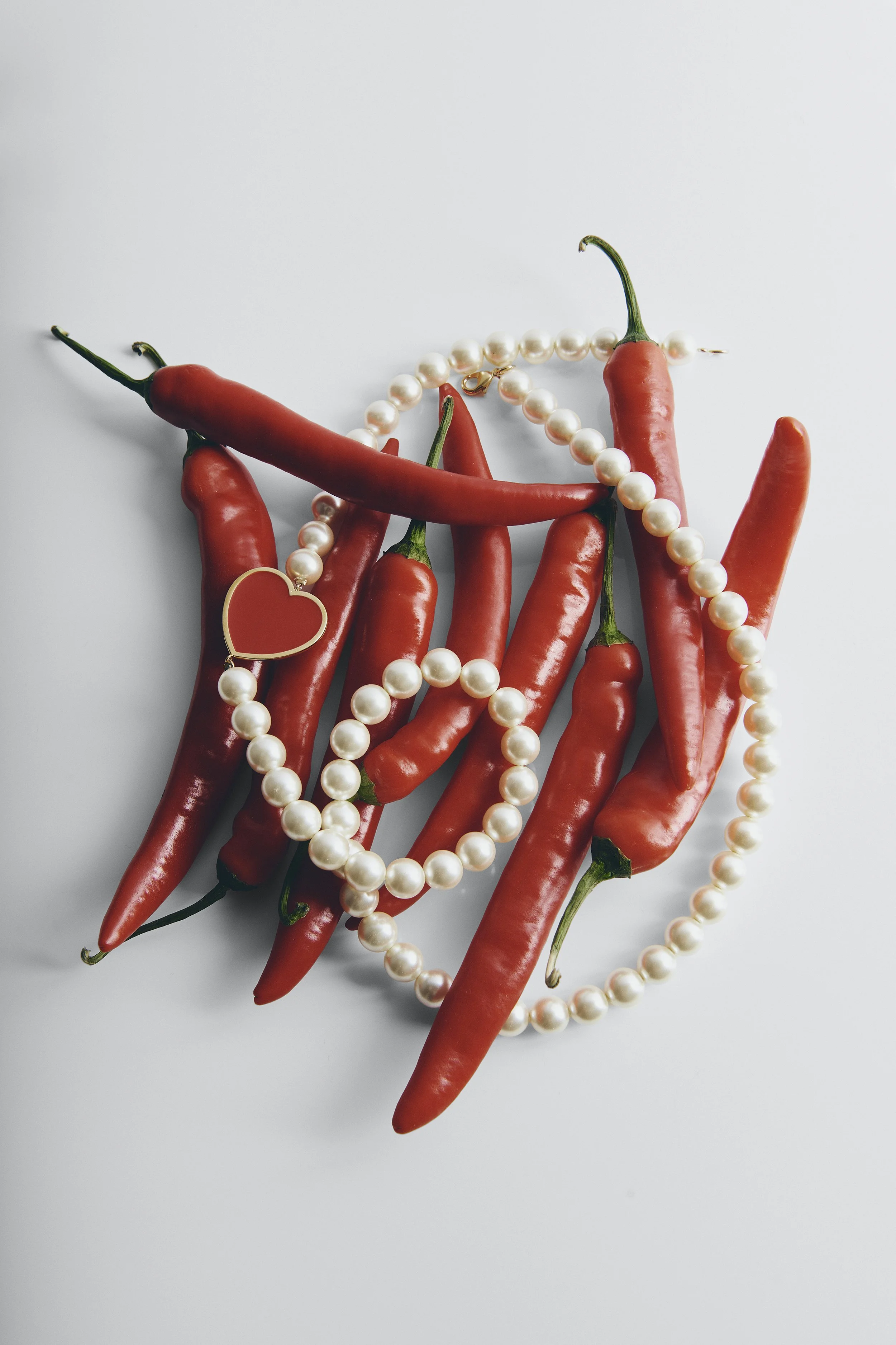 Red chili peppers intertwined with pearl necklaces and a small heart-shaped jewelry pendant on a white background.