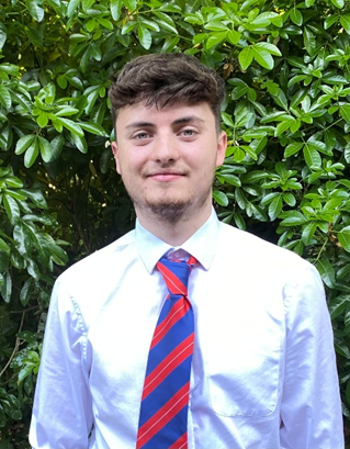 Young man with short brown hair, wearing a white dress shirt and a red and blue striped tie, standing in front of green leafy bushes.