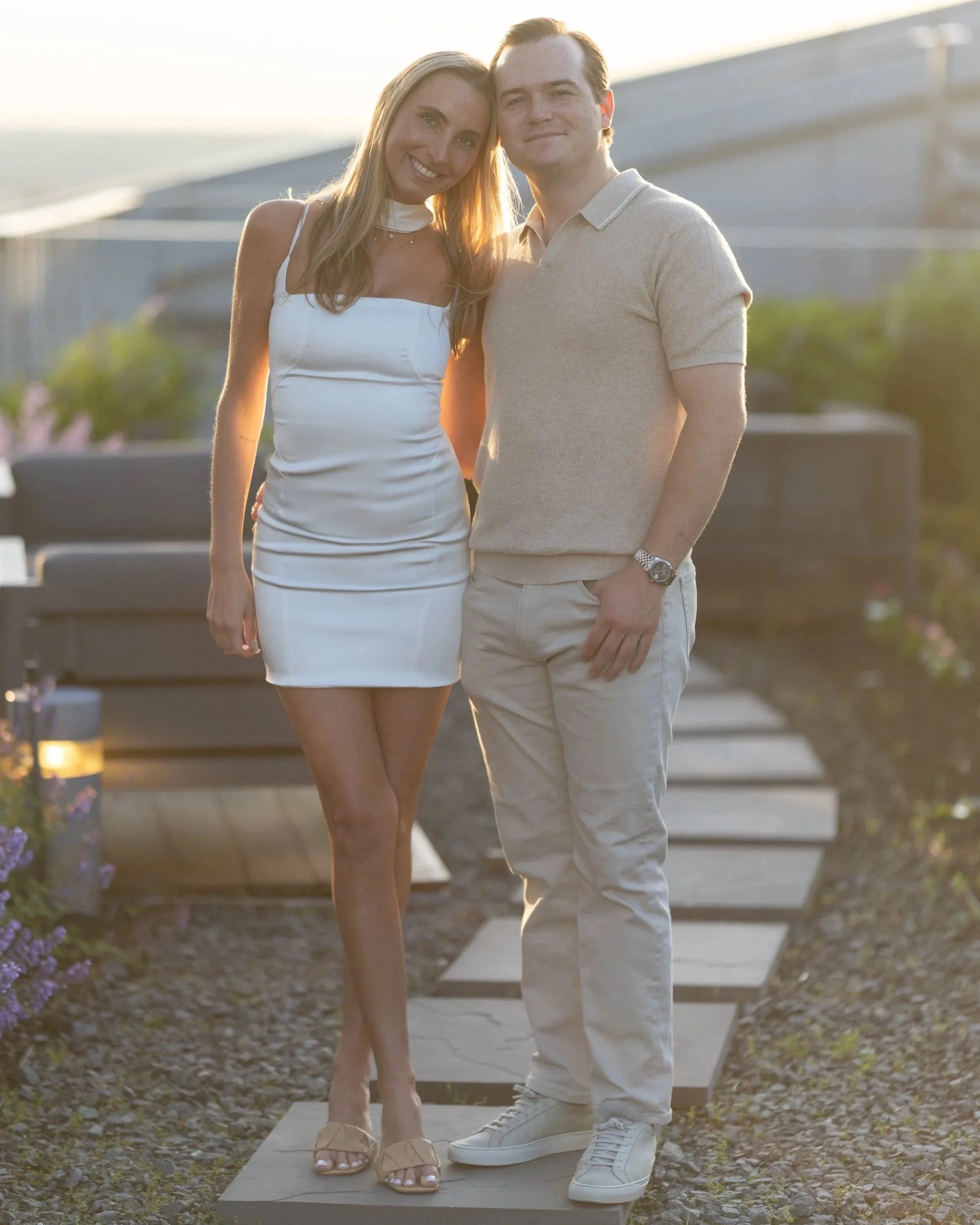 A smiling woman and man stand close together outdoors during sunset, with a staircase in the background, dressed casually in light-colored clothing.