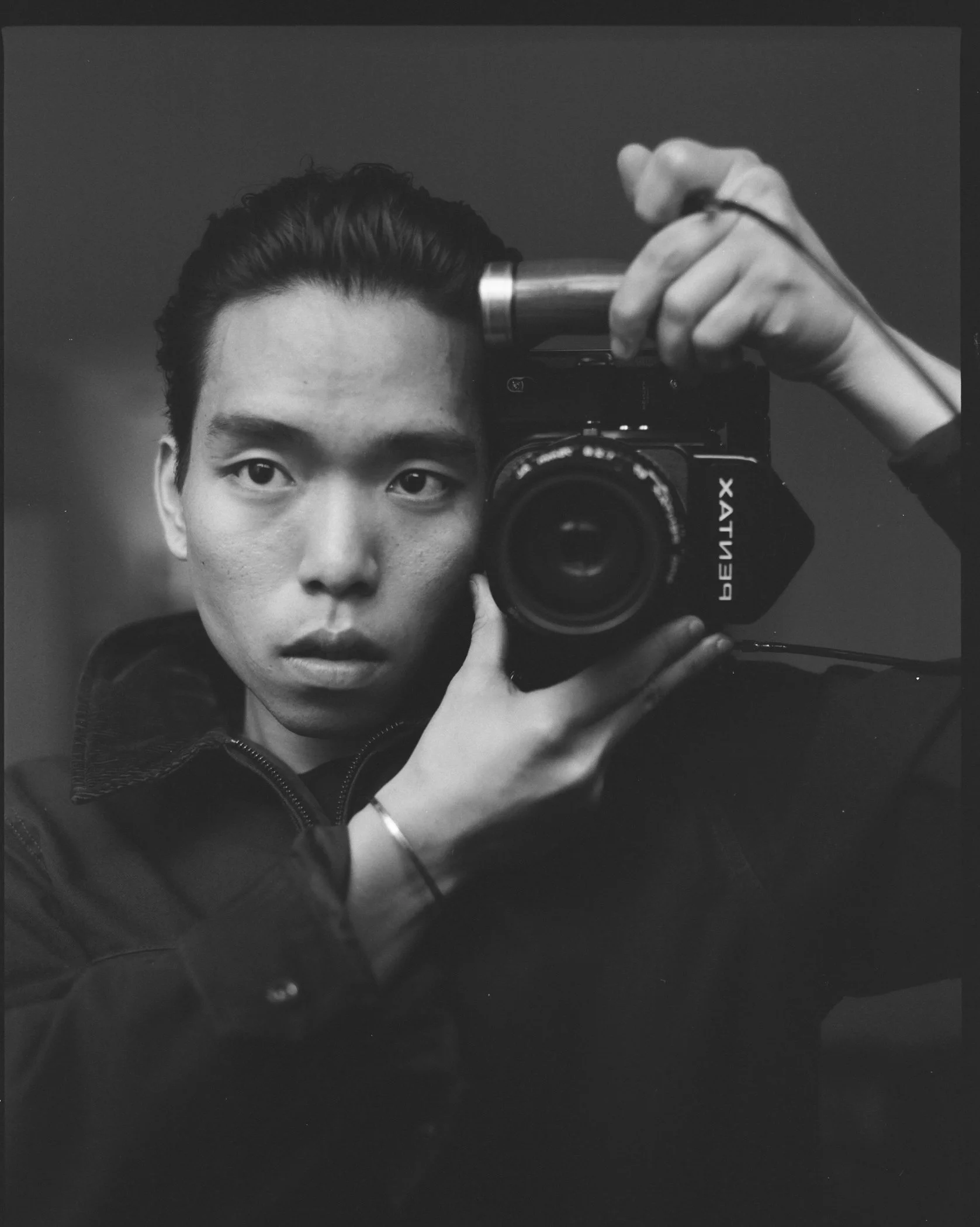 Black and white photograph of a person taking a mirror selfie with a digital camera, their face partially visible and hand holding the camera in front of their face.