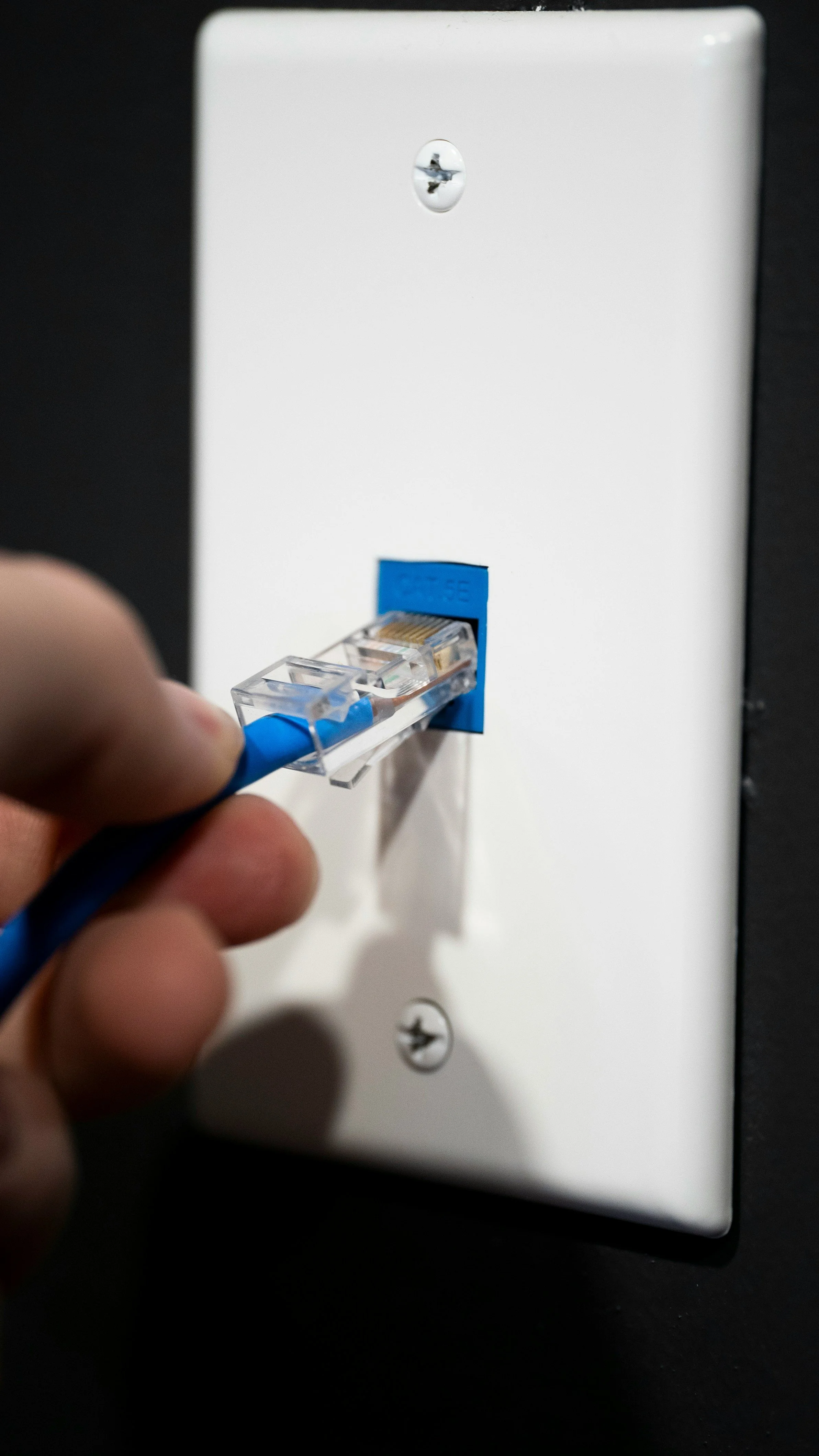 Close-up of a person's hand plugging a blue Ethernet cable into a wall jack.