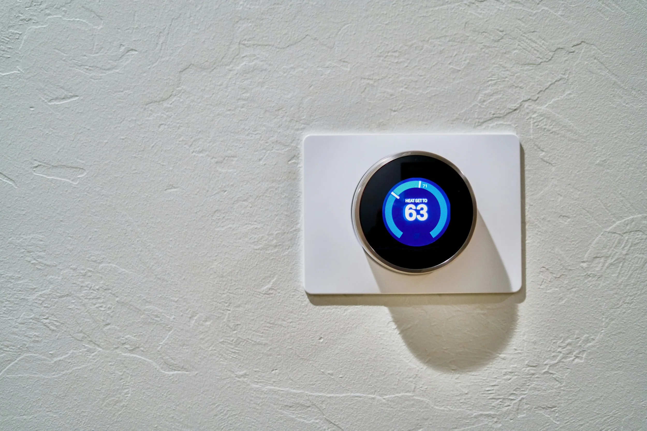 Smart thermostat mounted on a wall displaying a temperature setting of 63 degrees with a blue circular interface.