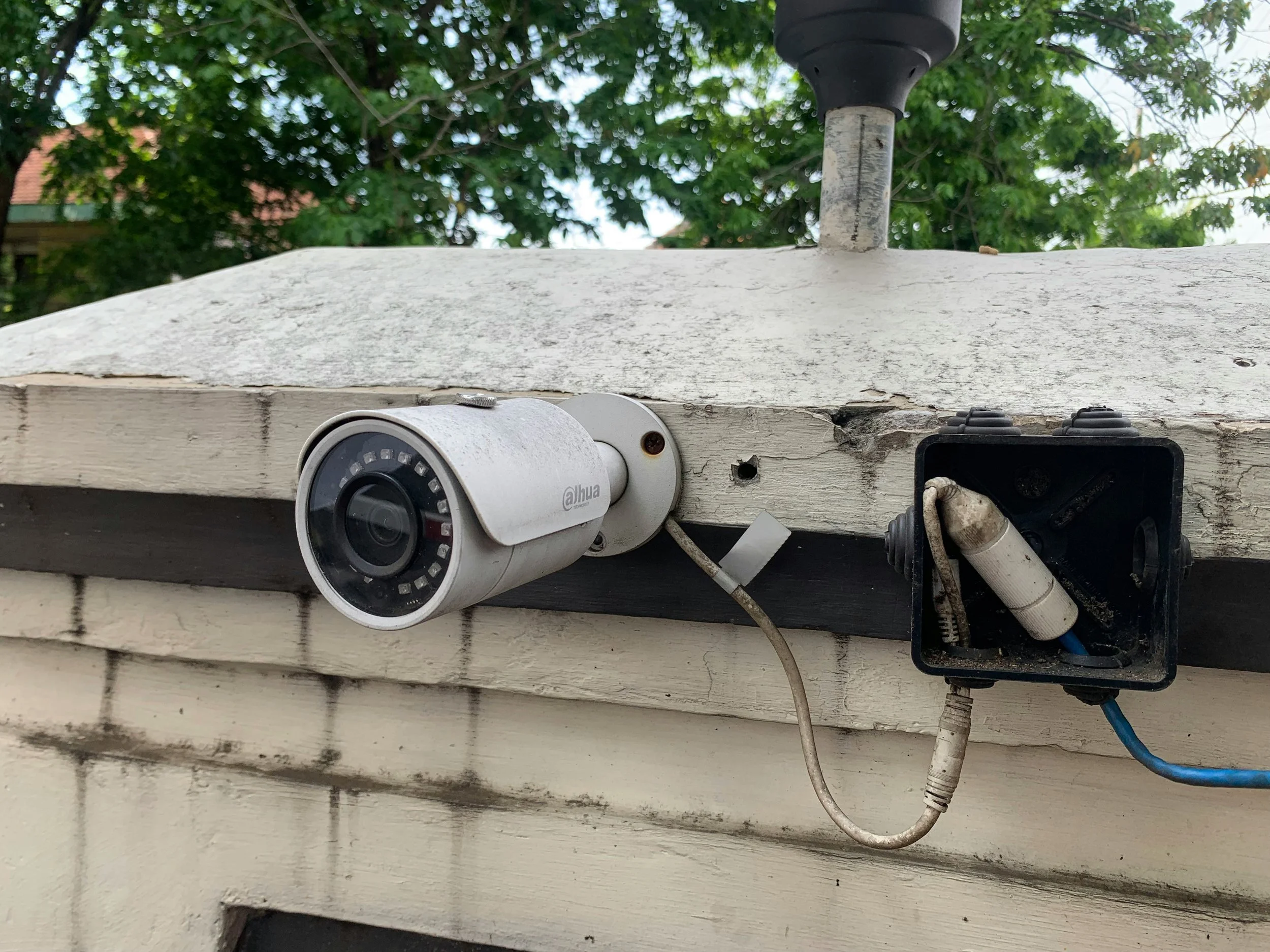 A security camera and an exposed electrical box mounted on the exterior of a building with peeling paint, with trees and a roof in the background.