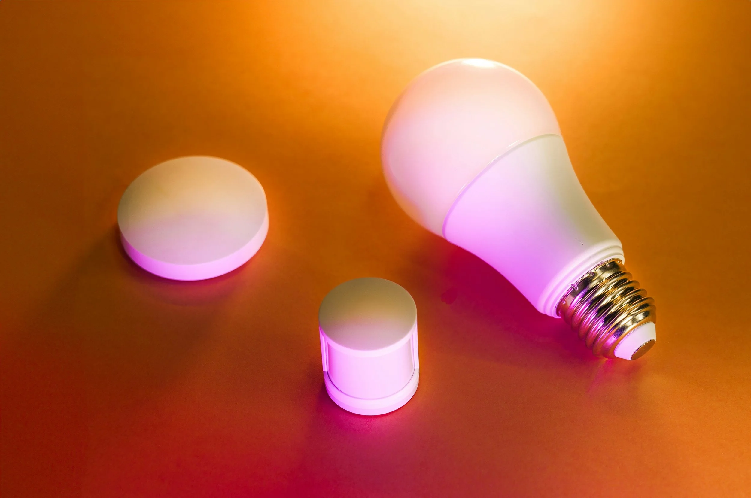 Smart light bulb with attached color-changing cap and two additional caps on a gold-colored surface, emitting pink light.