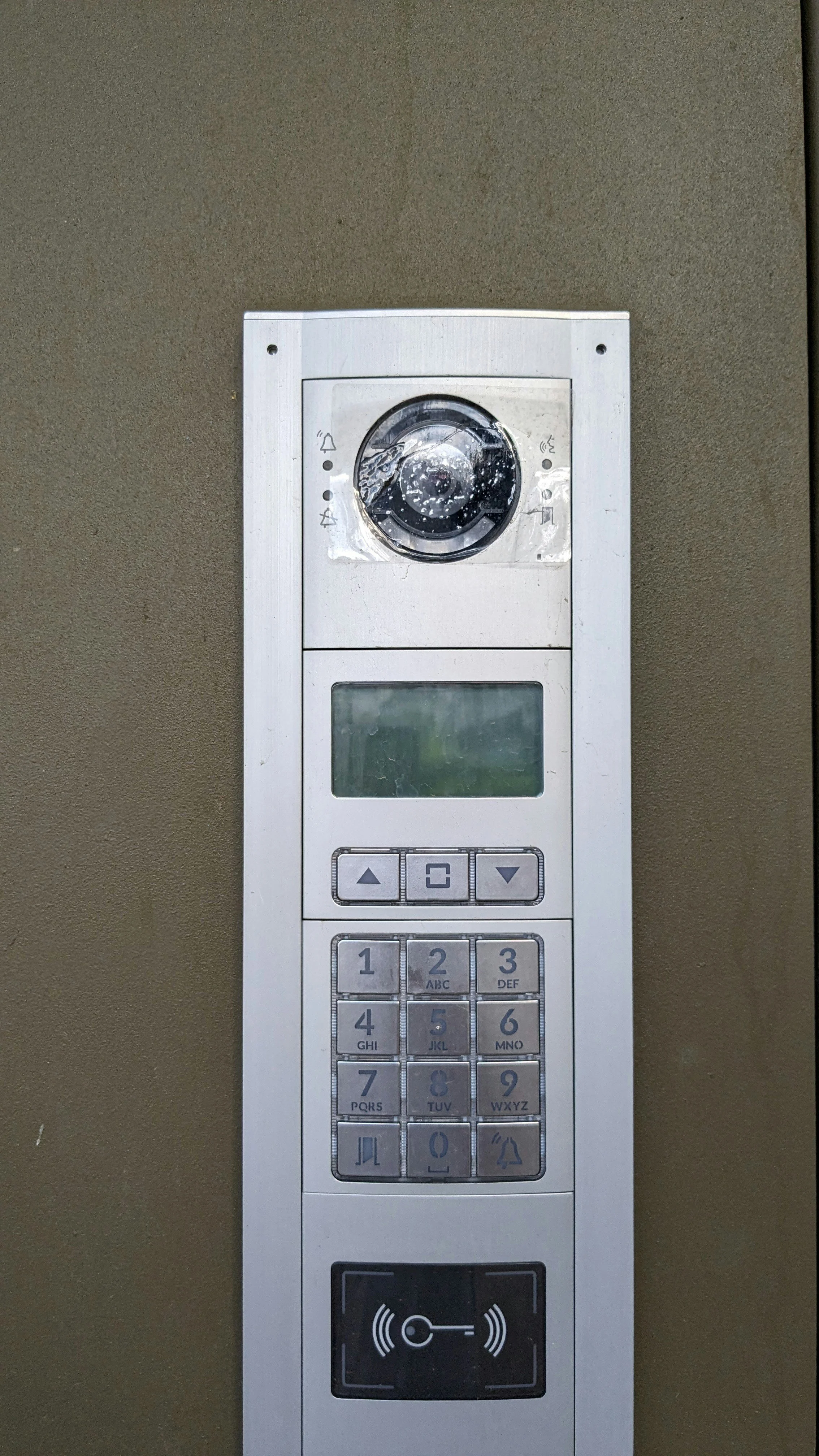 Office building intercom with keypad, screen, and speaker, mounted on a beige wall.