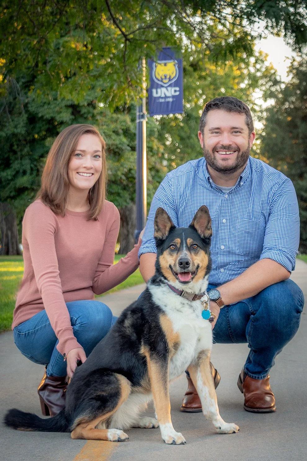 A smiling young woman, a smiling young man, and a happy dog sitting on a paved path in a park with green trees and a University of Northern Colorado flag in the background.