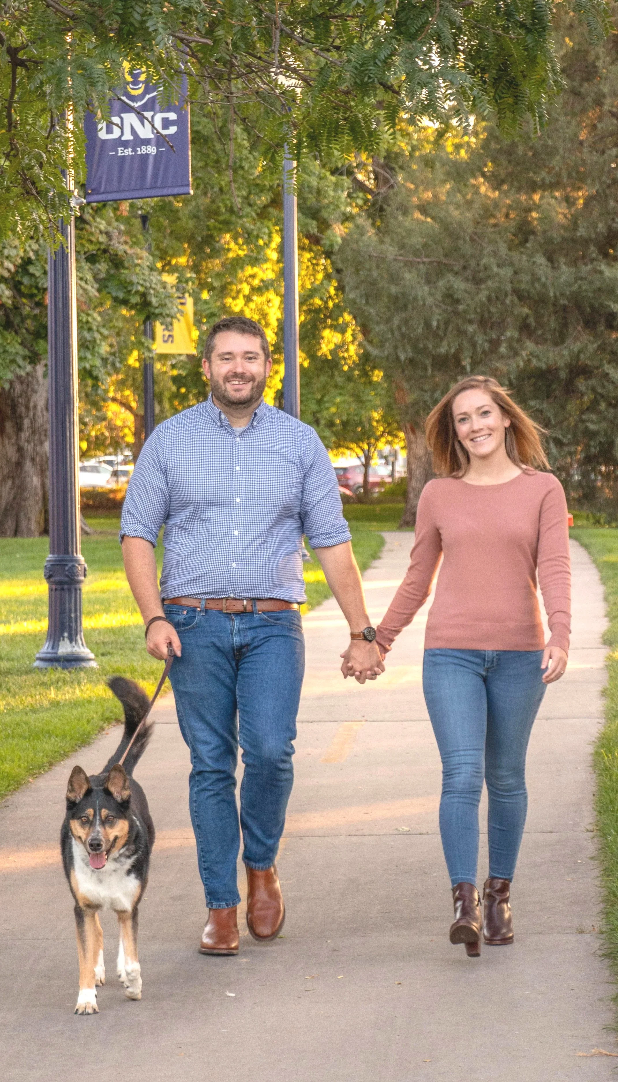 A young couple holding hands and walking their dog on a sidewalk in a park during sunset.