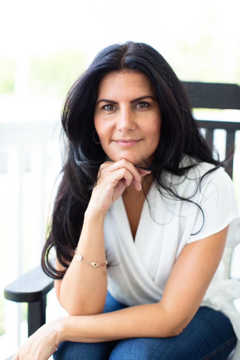 A woman with long dark hair and light skin sitting outdoors, looking at the camera with her chin resting on her hand, wearing a white top and bracelet, with a blurred background of a porch or patio.