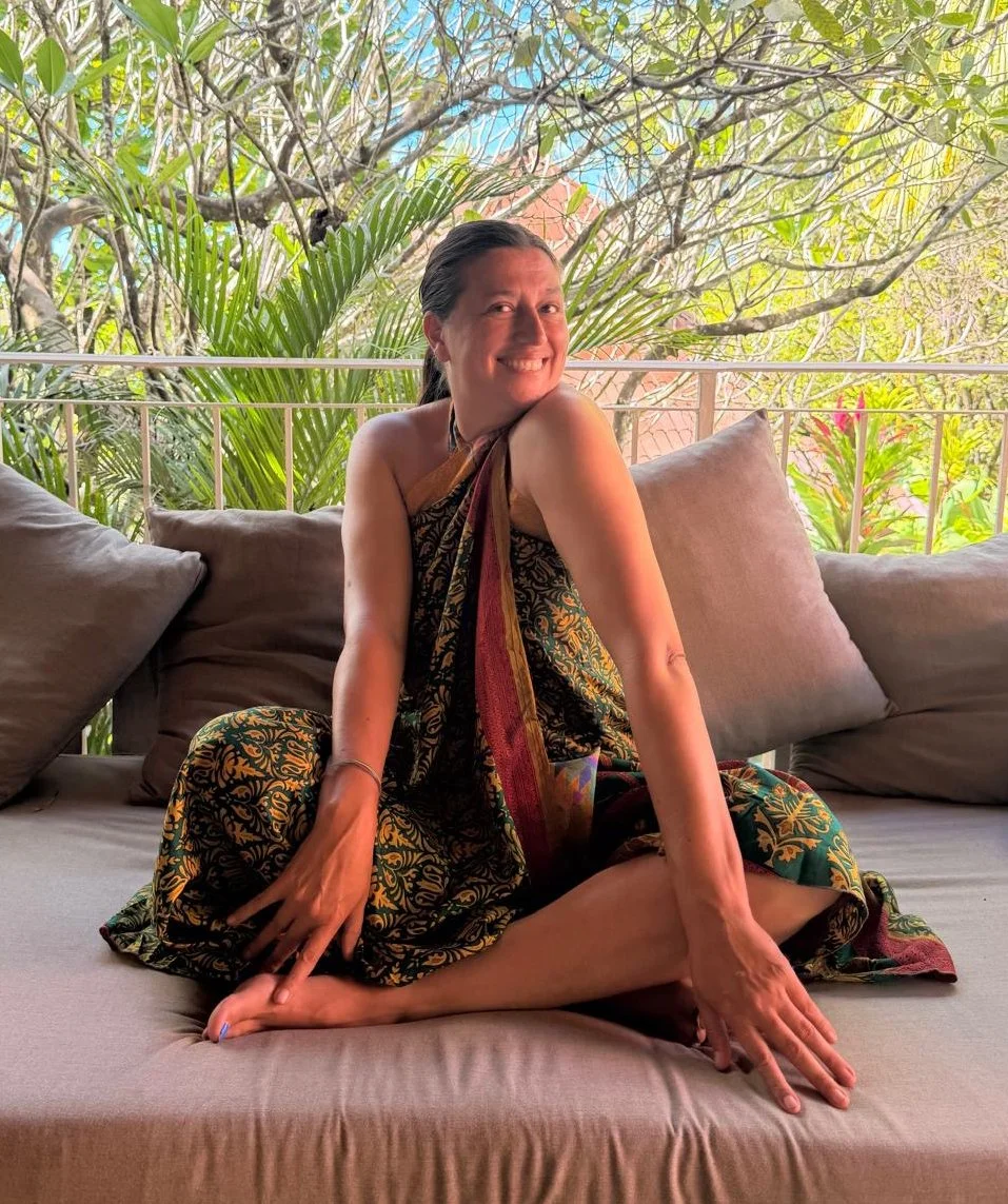 A woman sitting on an outdoor couch, smiling, with lush green trees and plants in the background.