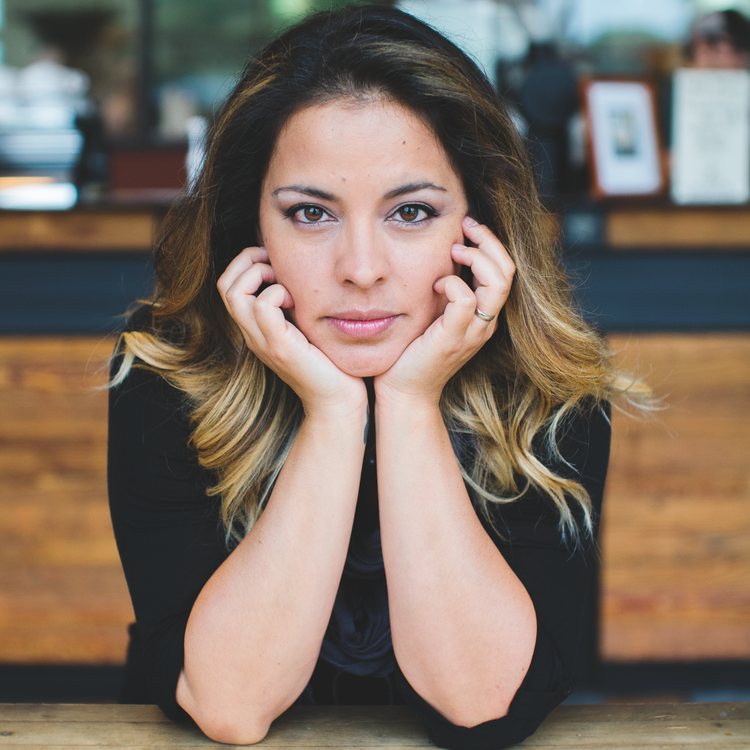 Woman with shoulder-length hair, looking directly at the camera with her face resting in her hands in a cafe or restaurant setting.