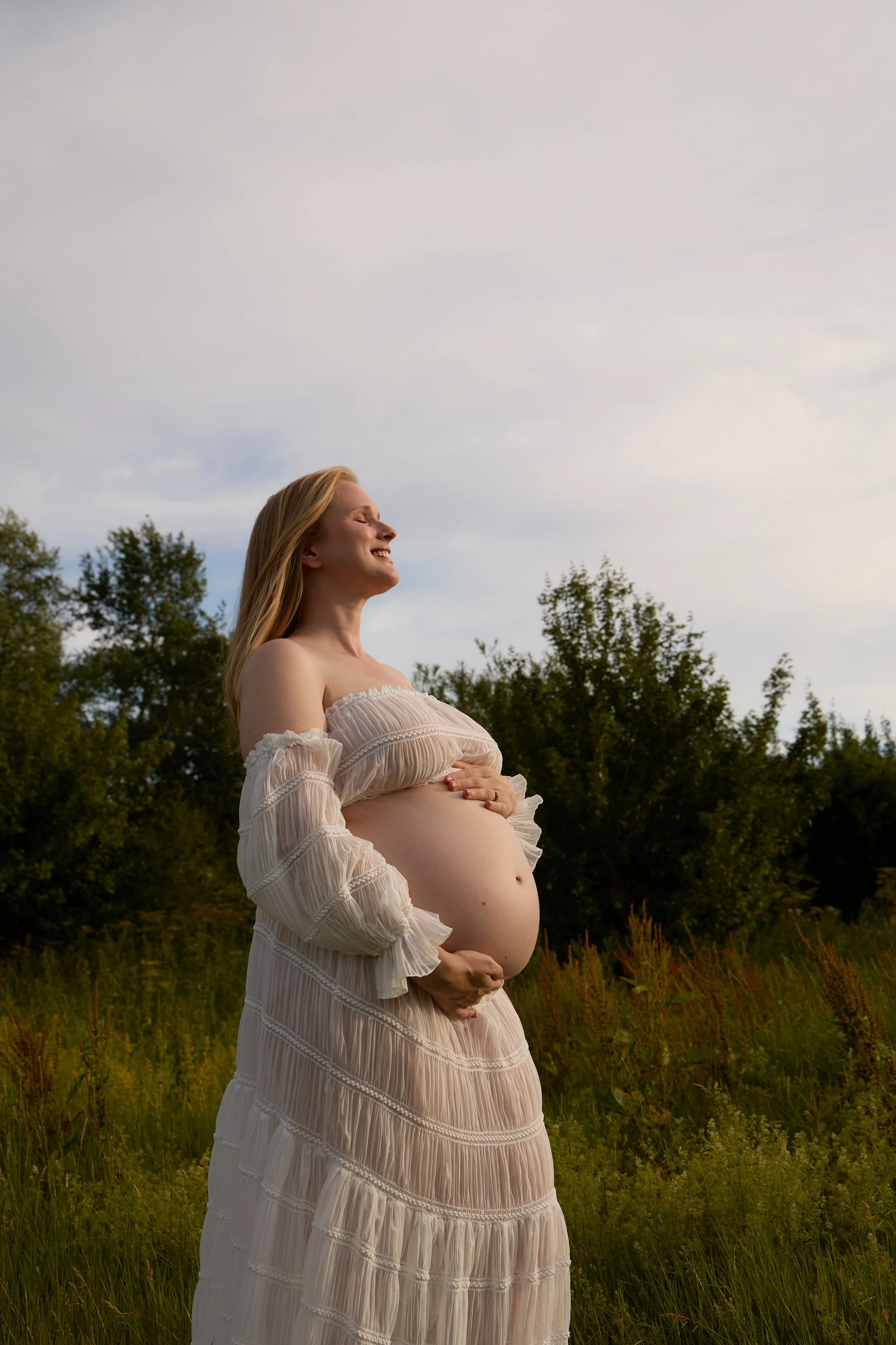 A pregnant woman with long blonde hair standing outdoors in a field, smiling with her eyes closed, holding her belly. Maternity photoshoot in Bicester, Oxford.