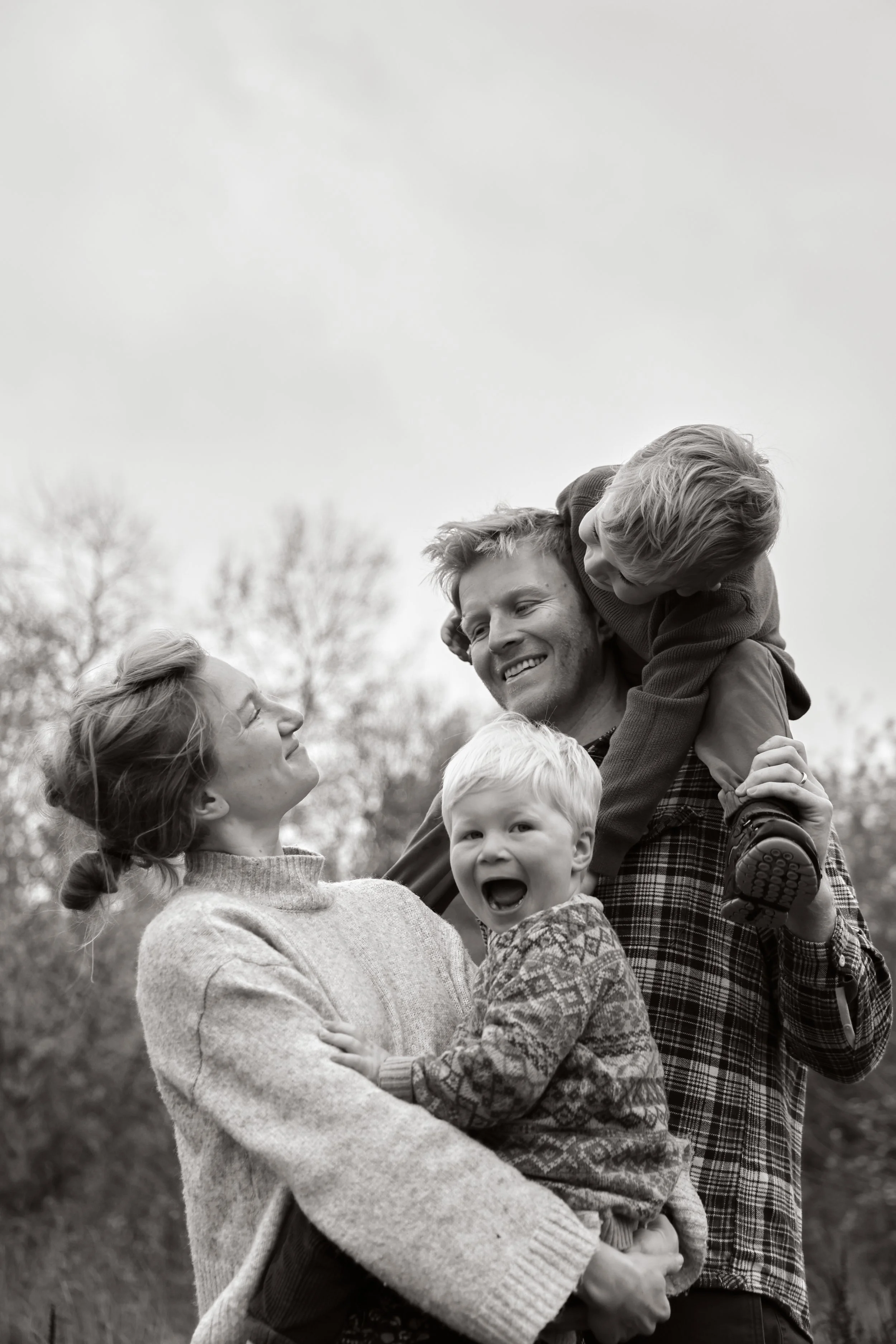 A happy family of five outdoors enjoying time together in black and white. The father is holding a young boy on his shoulders, and the mother is holding a younger boy. The family is smiling and interacting affectionately.