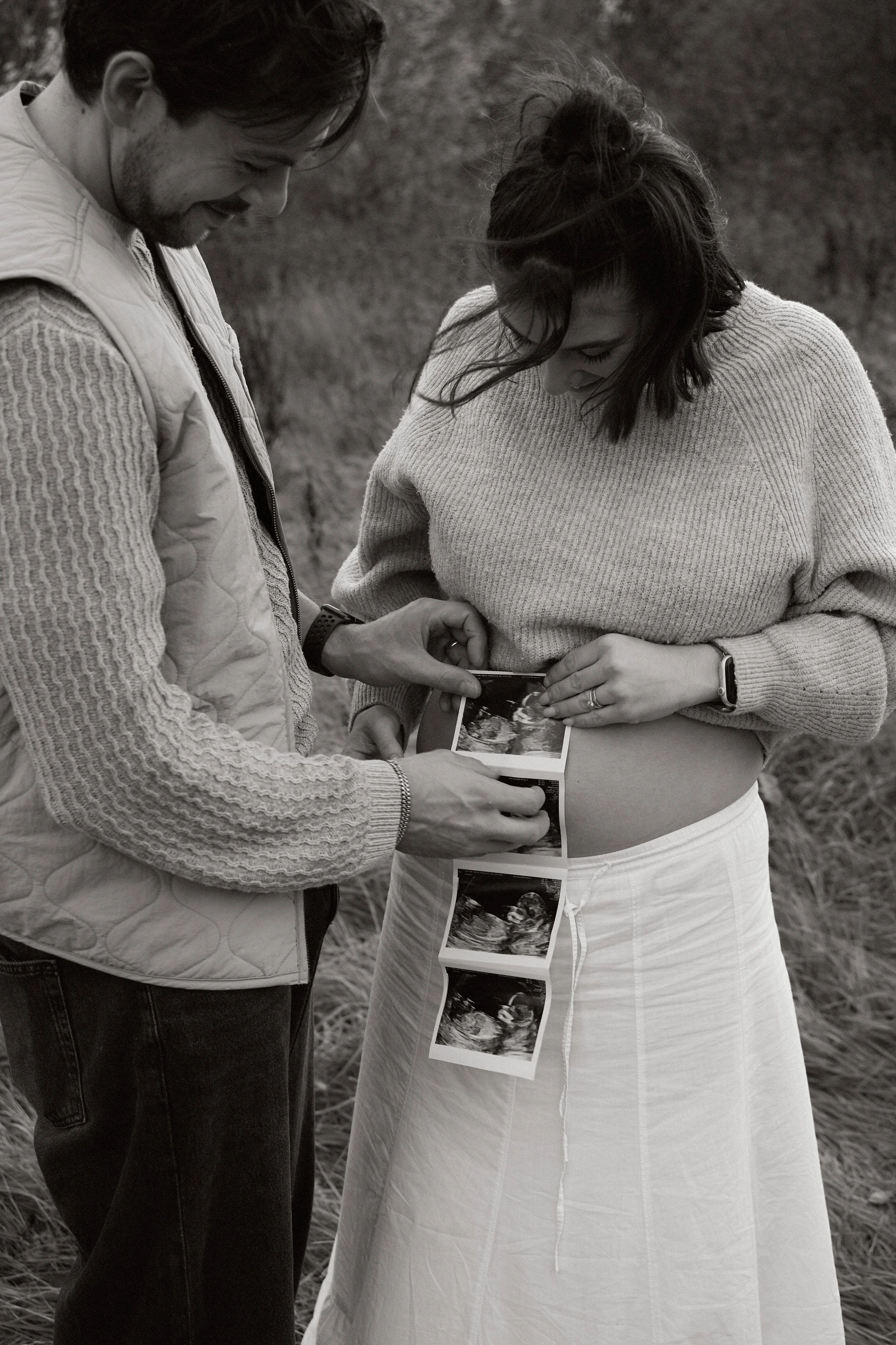 A pregnant woman and a man looking at ultrasound photos together outdoors. Maternity photoshoot in Bicester, outside, natural, black and white.