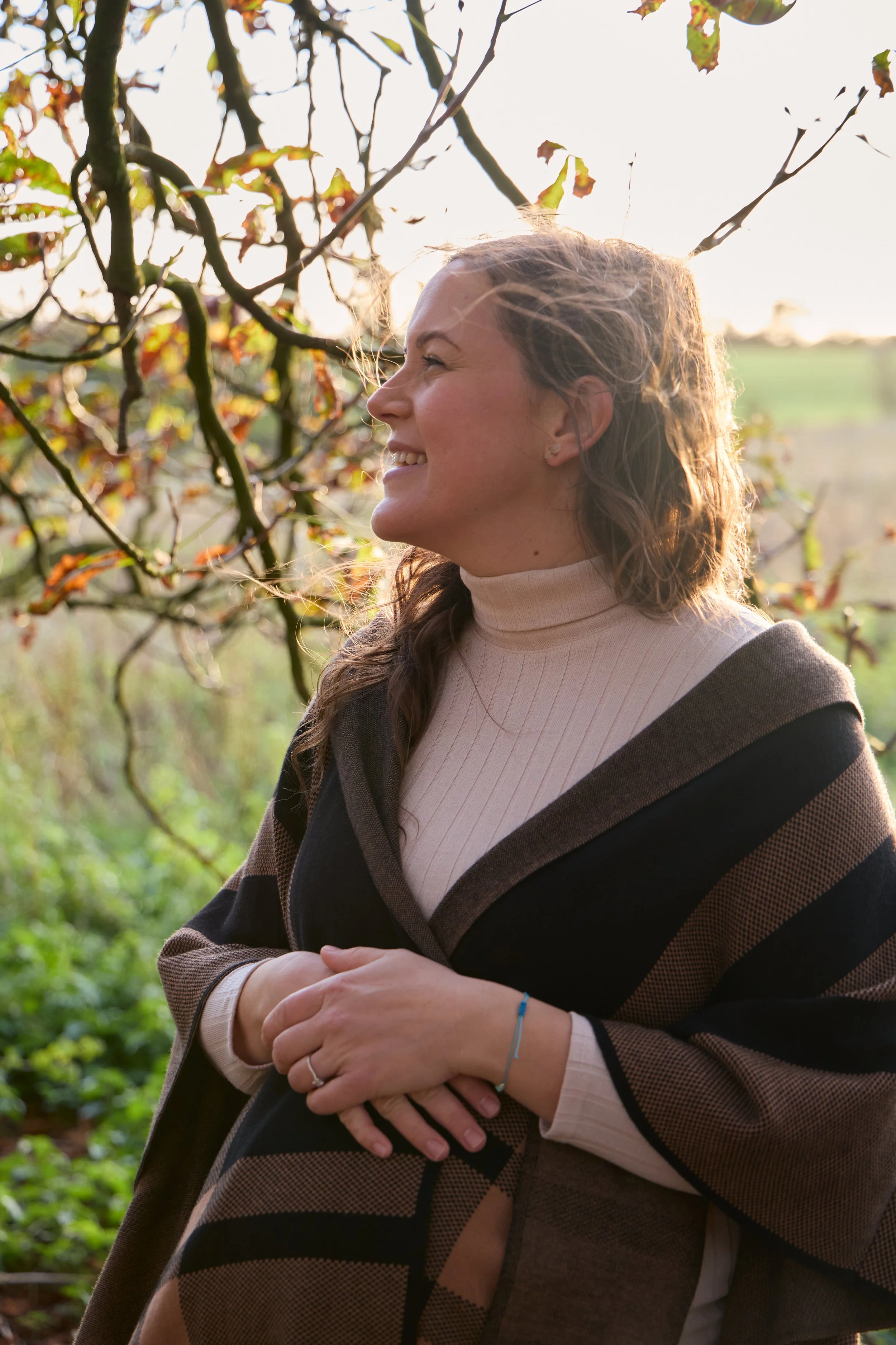 A woman standing outdoors in front of a tree with falling leaves, smiling and looking to the side, during late afternoon or early evening. Maternity photoshoot in Bicester outside.